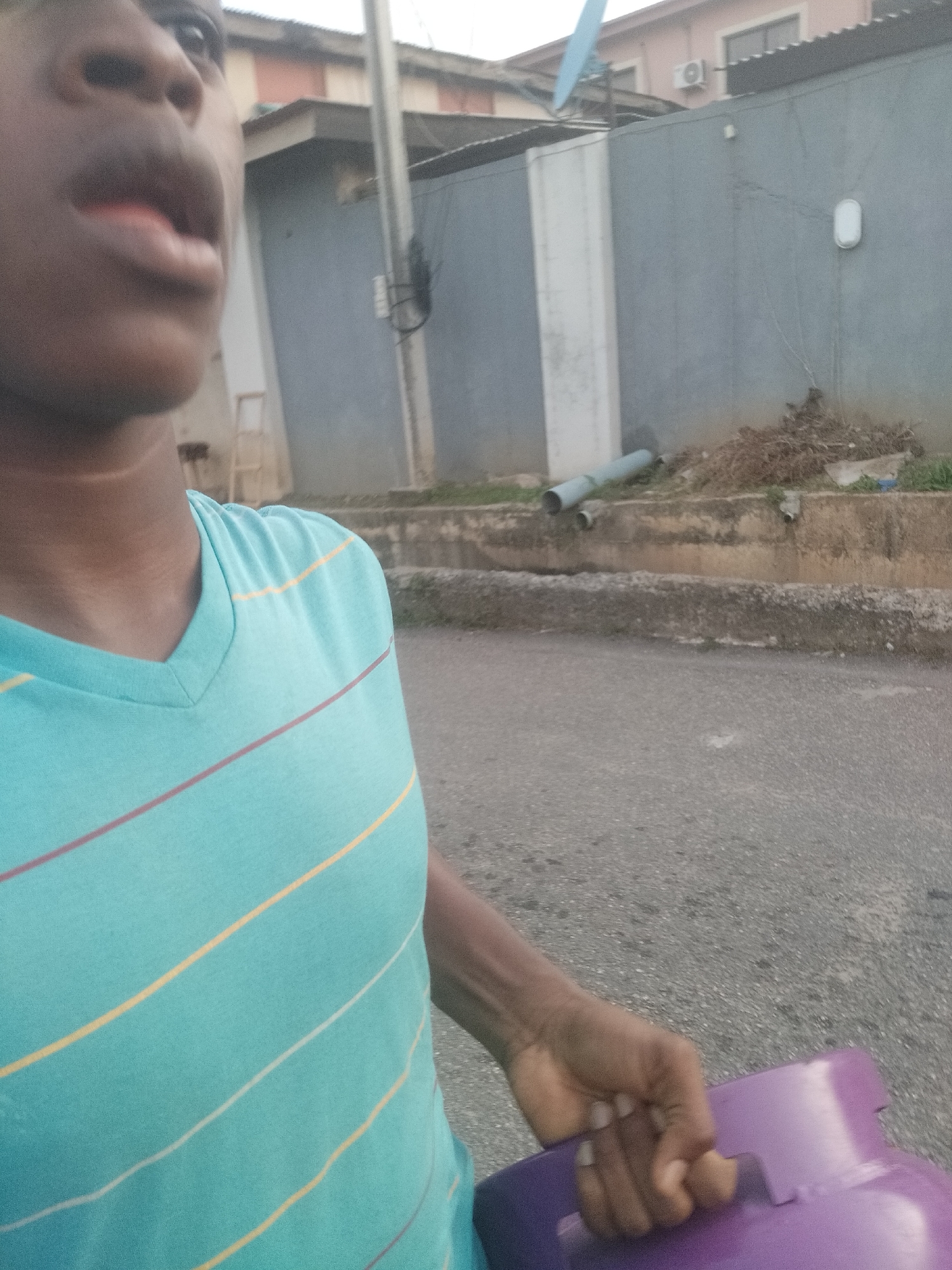 The image shows a person of African descent, possibly male, holding a purple container with a handle. The person is wearing a turquoise, V-neck t-shirt with horizontal yellow, white, and red stripes. The background suggests an outdoor location in Shangisha, Nigeria. There is a paved road and a wall with a light fixture. Pipes are visible along the base of the wall. In the background, there is a building with what appears to be a satellite dish attached to it. A utility pole is also visible.