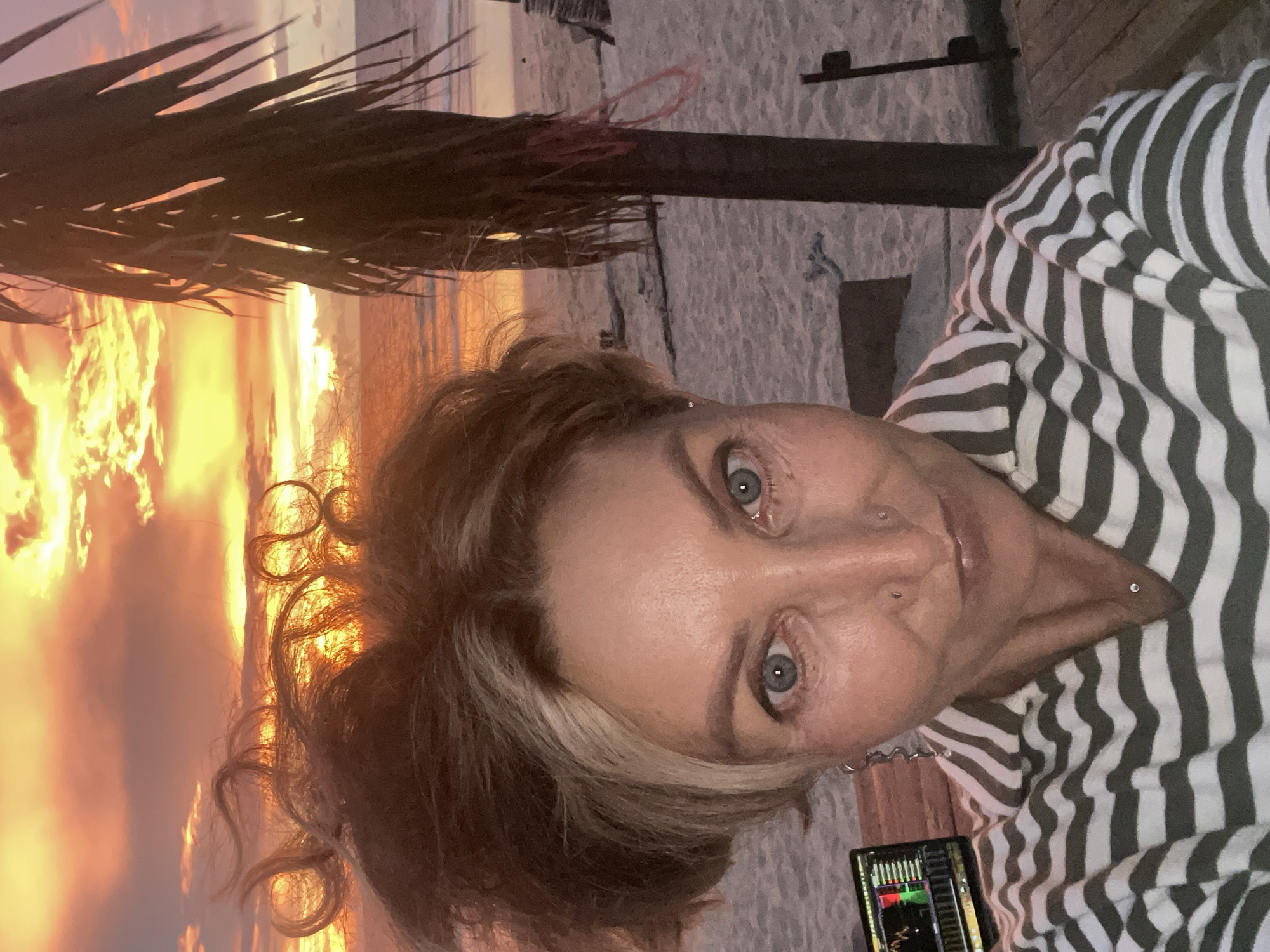 A woman is taking a selfie on a beach in Choeng Thale, Thailand, during a sunset. The sky is ablaze with orange and yellow hues from the setting sun, and clouds are scattered across the horizon. A palm tree trunk with some attached pink rope is visible on the right side of the frame. The woman has light blue eyes, blonde and brown hair styled in loose curls, and a nose ring. She is wearing a striped shirt. A laptop displaying a financial chart is positioned to her left. The sand is visible in the foreground, with some wooden structures and debris scattered on it. The ocean is visible in the background.