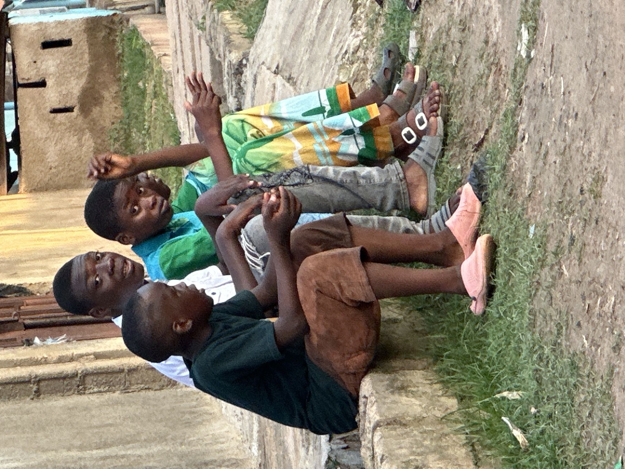 Three young Black males are seated on a concrete ledge outdoors, with grass and dirt in the foreground. The person on the left wears a dark green t-shirt and brown shorts. The person in the middle wears a light blue shirt and grey pants. The person on the right wears a green shirt with a colorful, patterned fabric draped over their lap. Their footwear is visible: the person on the left wears pink slides, the person in the middle wears grey slide sandals, and the person on the right wears dark sandals. The background consists of a textured, light-colored wall with dark openings and hints of a blue structure to the right. The scene appears to be in Narayi, Nigeria.