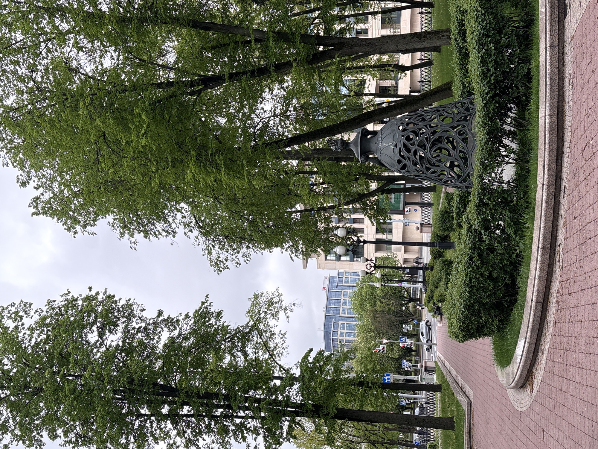 The image shows a park in Minsk, Belarus. In the foreground are trees with light green leaves and a brick sidewalk edged with a concrete curb. Adjacent to the sidewalk is a hedge surrounding a decorative metal structure. Further down the sidewalk, there are street lamps, traffic signs, and cars visible in the distance. Buildings can be seen in the background. The sky is overcast.