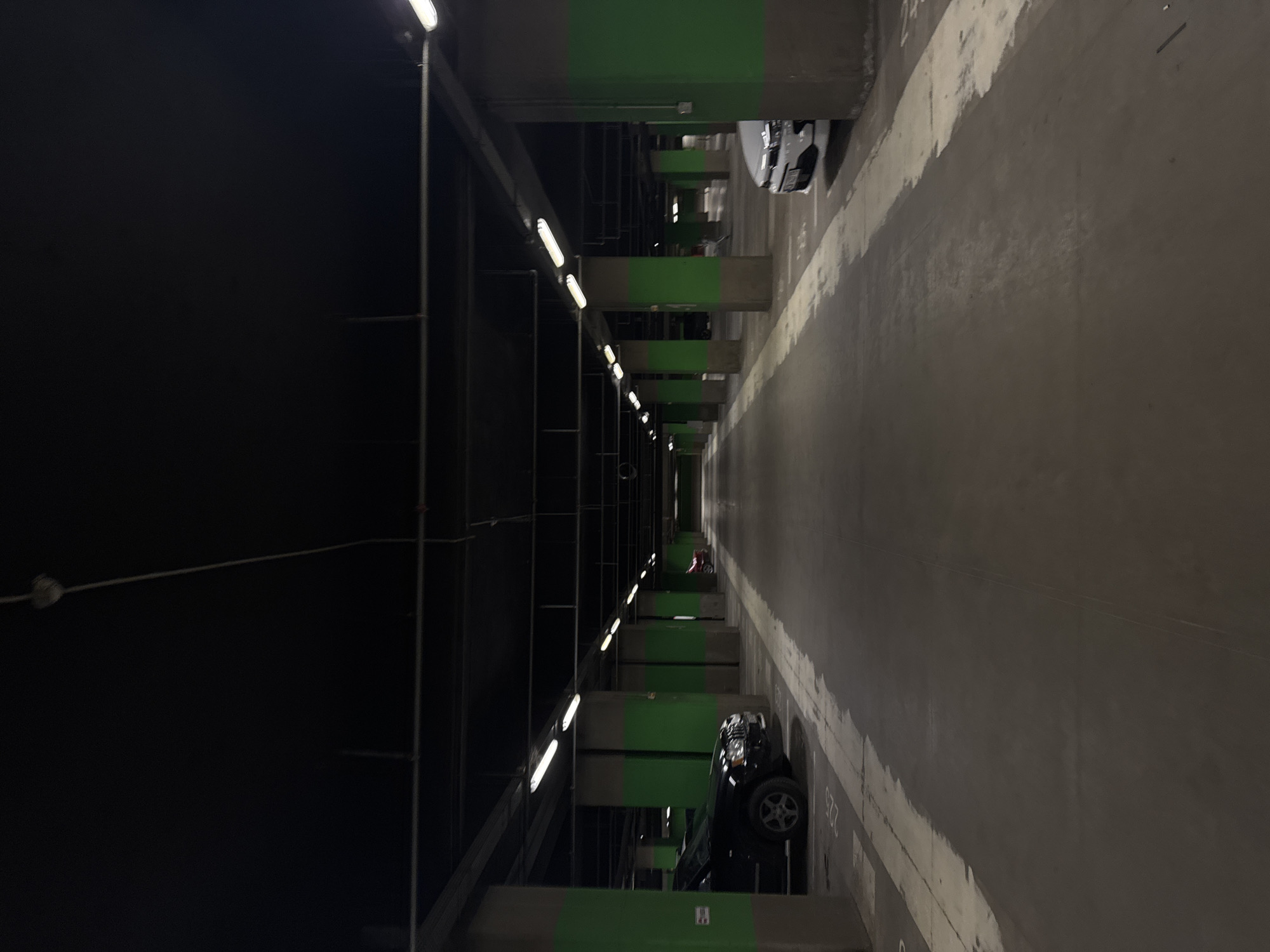 The image shows the interior of a parking garage. The walls are grey and marked with white lines indicating parking spaces. A black car is parked on the right side of the frame, partially visible. In the distance, another vehicle, possibly a small red child's scooter or toy car, is seen. Overhead lighting fixtures illuminate the garage space. Structural pillars, painted with green accents, are located along the length of the garage. The left side of the frame is darker, revealing ventilation pipes and electrical fixtures. The location is Sofia, Bulgaria.