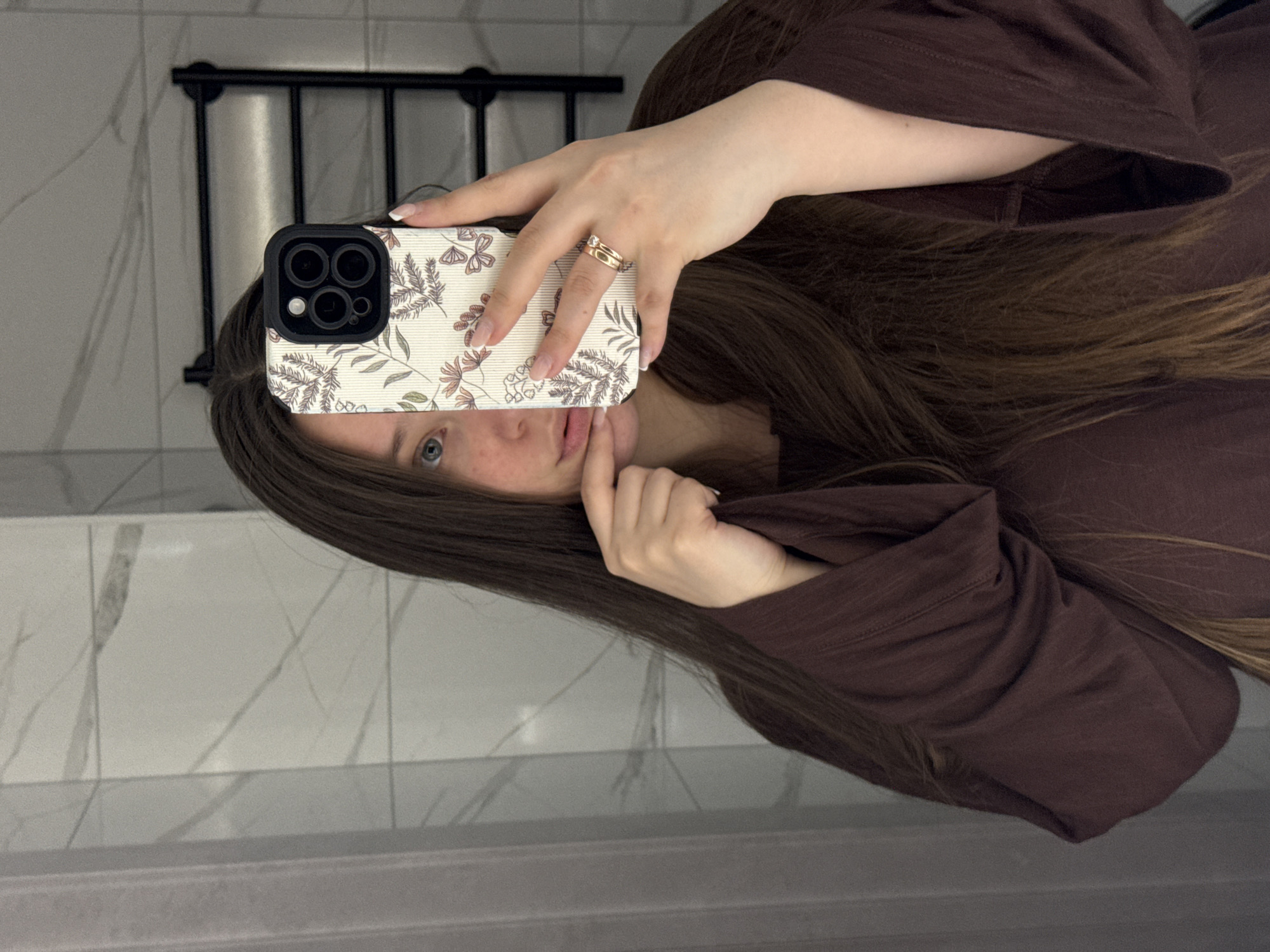 A female individual is depicted in a mirror selfie, partially obscuring her face with a smartphone. The subject has long, dark brown hair and wears a dark brown long-sleeved top. Her left hand holds an iPhone, equipped with a cream-colored case featuring a botanical pattern of leaves, flowers, and butterflies. The phone's multi-lens camera module is visible. The ring finger of the left hand displays a gold-toned ring. Her right hand is positioned near her chin, with fingers touching the lower lip. The subject's right eye, appearing blue-green, and a portion of her forehead, nose, and lips are visible. In the background, white and grey marble-patterned tiles cover the wall, with a black, multi-tiered towel warmer mounted on the right. A light-colored curtain or fabric is partially visible on the left side. The scene is an interior room, consistent with a bathroom, located in Krasnodar, Russia.