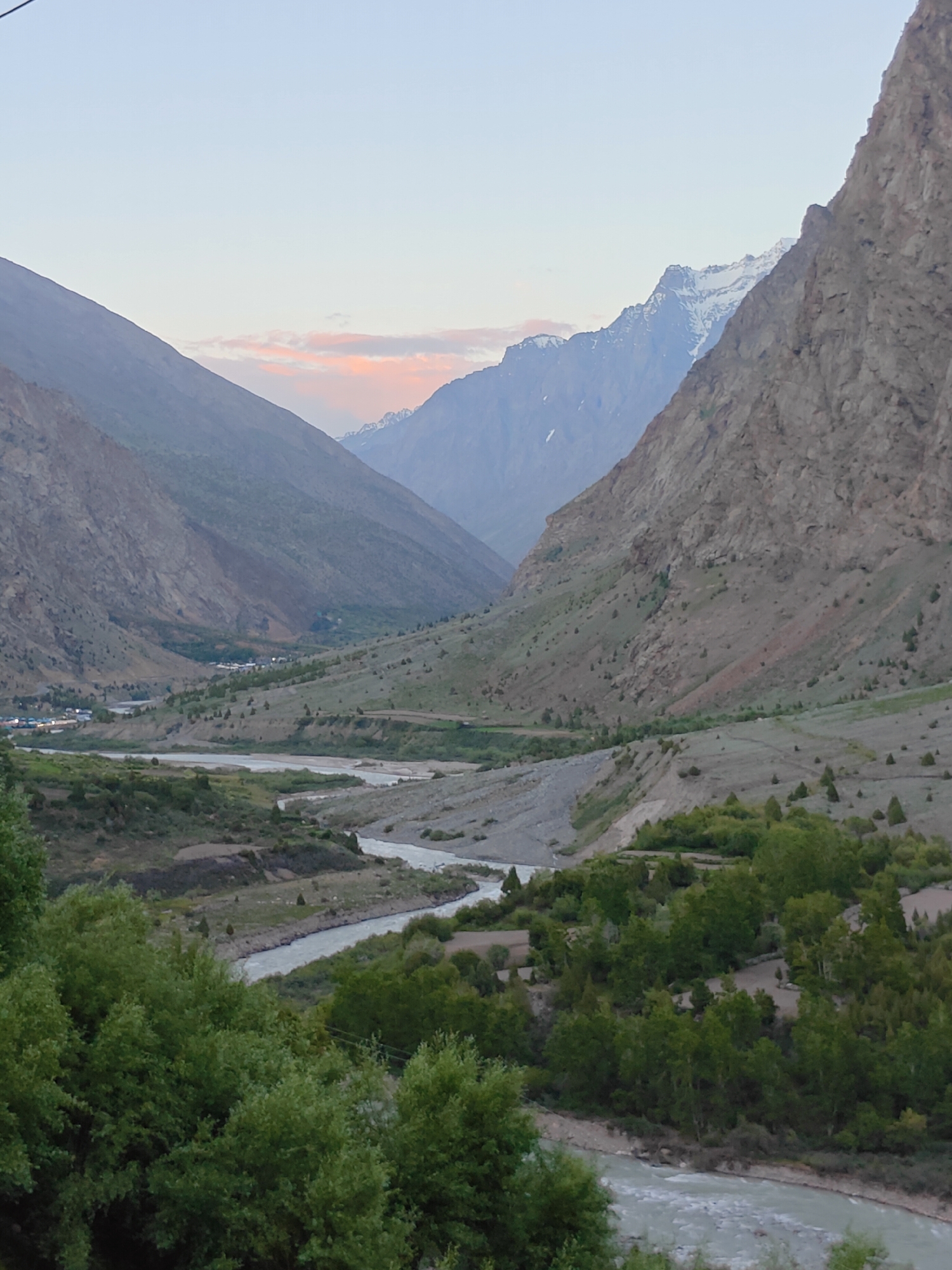 The image depicts a serene valley landscape in Gemur, India, during the twilight hours. The sky is a pale blue with a hint of soft pink and orange clouds, suggesting either sunrise or sunset. Dominating the foreground is a lush green canopy of trees, with their leaves appearing slightly blurred due to motion or depth of field. Beyond the trees, a meandering river flows through the valley, its water appearing a light grey or silver.
The middle ground reveals a vast, undulating terrain characterized by terraced fields and sparse vegetation, including scattered trees and bushes. The slopes of the valley are covered with a mixture of dry grass and rocky outcrops. In the distance, a small village is nestled within the valley, with clusters of buildings and lights visible.
The background is defined by imposing, majestic mountains. The slopes of these mountains are steep and rocky, with some peaks showing patches of snow. The overall atmosphere is tranquil and natural, showcasing the raw beauty of the Himalayan region. The scene is devoid of any people or animals.