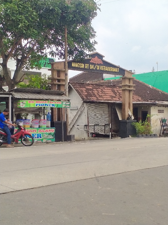 This street-level photograph captures a community entrance in Kecamatan Kebakkramat, Indonesia, under an overcast sky during daytime. A prominent, ornate gate spans a path leading into the neighborhood, featuring a sign that reads "NANGSRI RT 04/DI KEBAKKRAMAT." A metal barrier arm extends across this entrance.
To the left of the gate, a modest street vendor stall with a corrugated roof is set up, advertising "ES BUAH ALAMI PIONER" (Natural Fruit Ice PIONER). Several large, clear containers of drinks and water bottles are visible on the stall. A man in a blue shirt is seated on a red motorcycle, parked on the asphalt road directly in front of the vendor stall.
Behind the stall and to the left, a large tree partially obscures a white building, which appears to be a mosque, with some visible text suggesting "MASJID FATTAH SRI KIDUL." To the right of the entrance gate, a small house with a traditional red-tiled roof and a closed roller shutter stands. The main asphalt road occupies the foreground, creating a clear separation from the community entrance. The scene depicts a quiet, functional local setting.