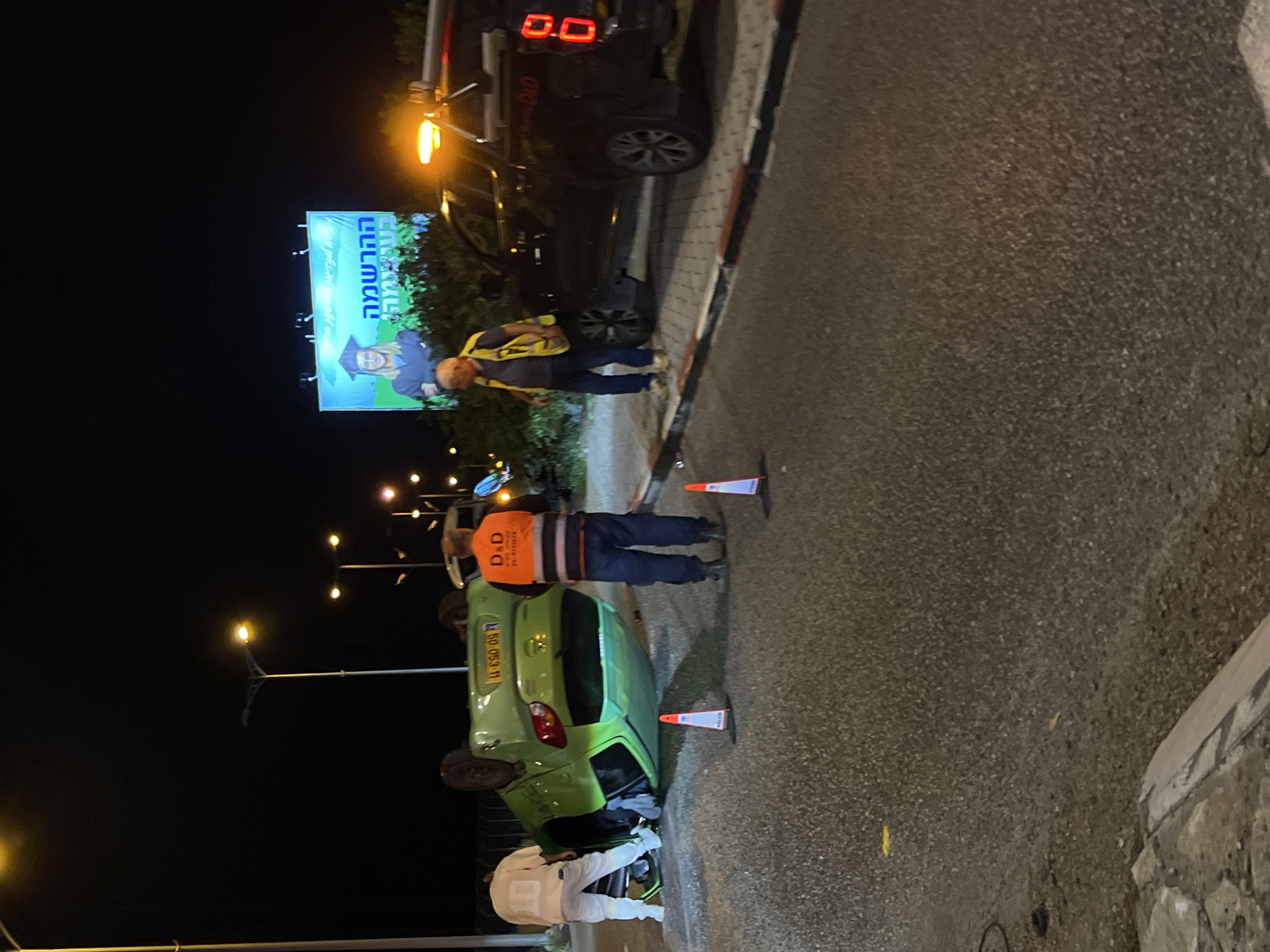 At night in Kiryat Motzkin, Israel, a green car lies overturned on the side of a road, surrounded by orange cones. Two men in reflective vests stand nearby: one is inspecting the overturned car, and the other is standing near a black tow truck. A billboard with an advertisement in Hebrew is visible in the background. A person in white clothing is near the car's wreckage. Streetlights illuminate the scene. The road is paved and bordered by a curb.