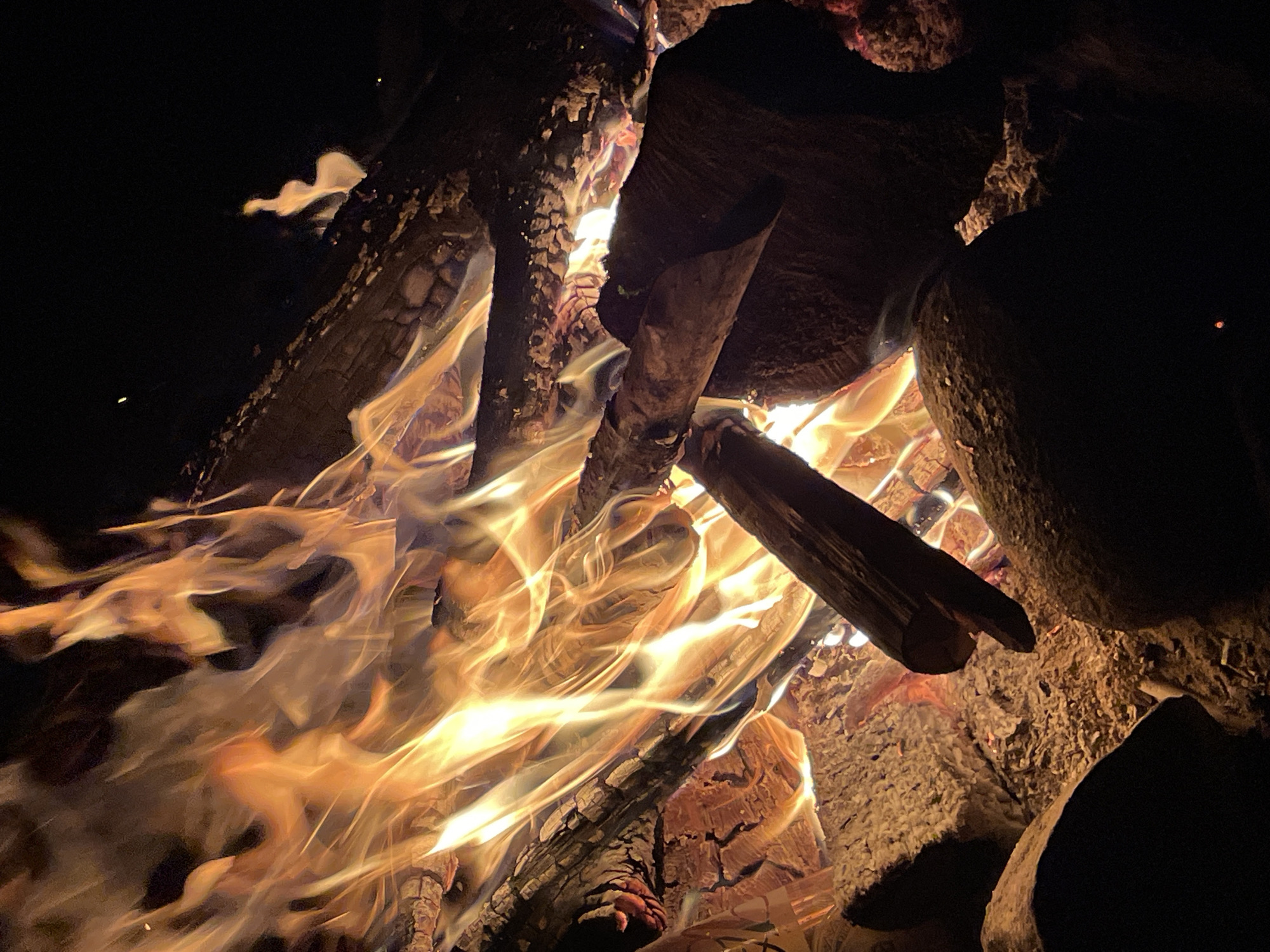 An outdoor bonfire burns brightly at night. The flames are a mix of orange and yellow, licking upwards and illuminating the surrounding logs and embers. Some of the logs are charred and cracked, indicating they have been burning for some time. The ground appears to be covered in ash and small stones. The background is dark, suggesting a rural or wilderness setting. There are no people visible in the image. The location in the city of Berozovo, Russia is not identifiable from the visual elements of the image alone.