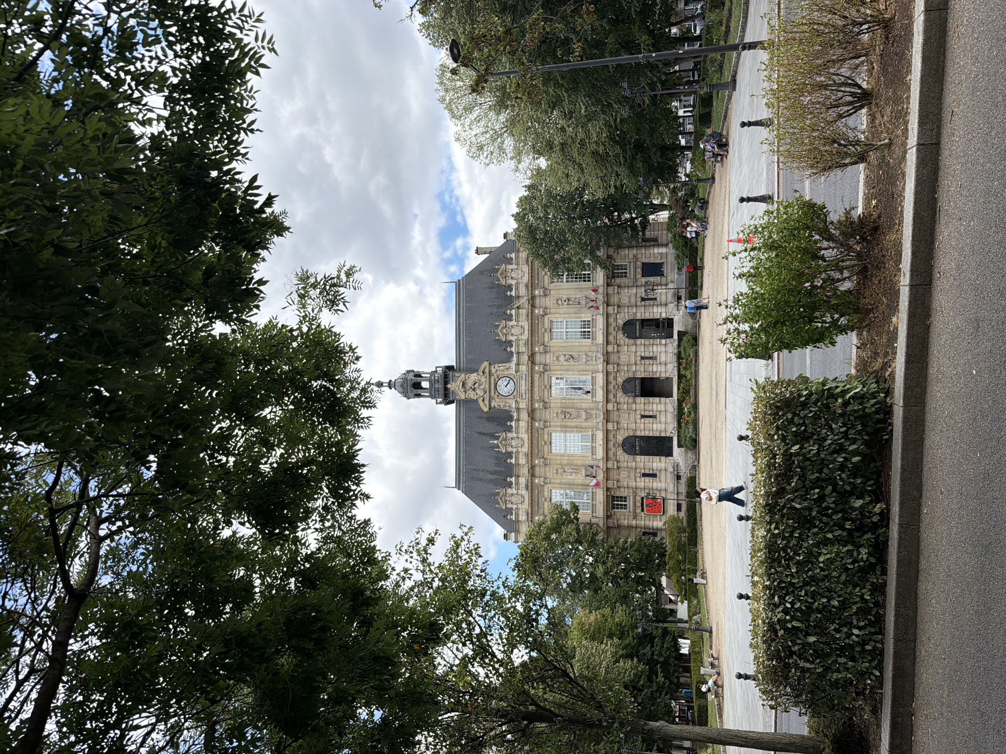 This media file shows a view of a building, possibly a town hall, with a clock tower. The building is made of stone with decorative architectural elements and multiple windows. Flags are visible on the facade. In front of the building, there is a paved walkway with benches and lampposts. Trees and hedges line the walkway and the foreground. Several people are seen walking or sitting on the benches, suggesting a public space.
The scene appears to be set in a town or city, with Ivry-sur-Seine, France, as the location context. The sky is cloudy with patches of blue, indicating a daytime scene with mixed weather. The overall mood is calm and public.
Visible text includes "ÉGALITÉ" written vertically on the building, and a red sign with a white symbol resembling a ladybug or a similar graphic. The clock on the tower is visible, but its hands are not clear enough to determine the exact time. The image is oriented vertically.