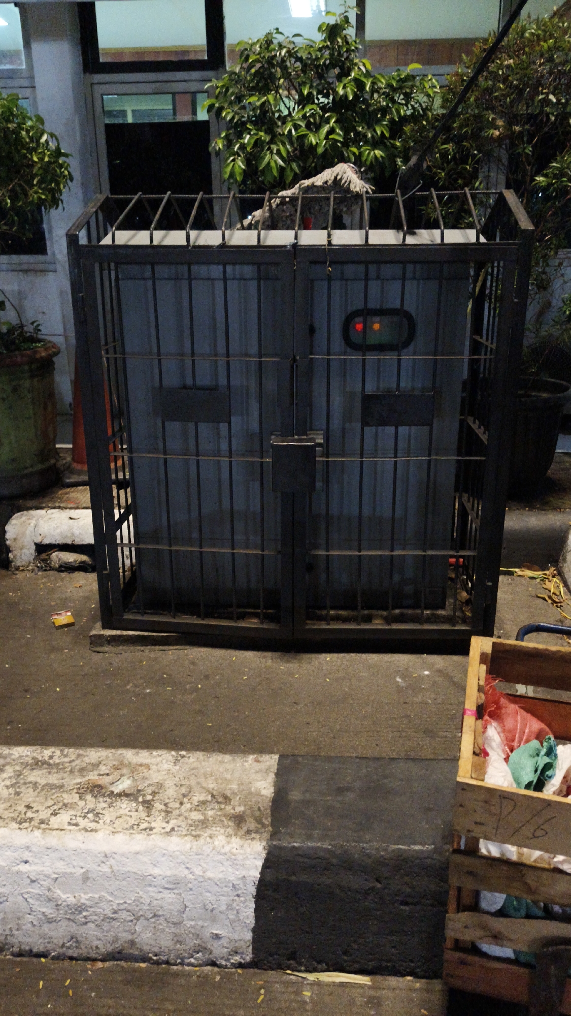 The image shows a caged trash receptacle located on a sidewalk in South Jakarta, Indonesia. The receptacle is made of metal bars and has a dark gray panel on its front. Two indicator lights are visible on the panel, one red and one yellow. The ground is paved with concrete and asphalt, with a painted curb marking the edge of the sidewalk. A wooden crate filled with what appears to be laundry or rags is situated to the right of the trash receptacle. In the background, there are plants and foliage, suggesting an outdoor urban environment. The overall scene depicts a typical street view in a city.