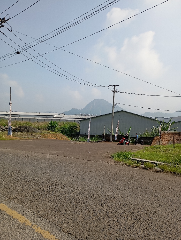 The image depicts an outdoor daytime scene in Pataruman, Indonesia. The foreground features a worn asphalt road with a faded yellow line on its left edge. To the right of the road, a narrow grassy verge transitions into an unpaved area. In the midground, a series of elongated, light-colored industrial or warehouse buildings with sloped roofs span horizontally across the frame. Multiple vertical banners are erected in front of these structures. A large dirt mound is visible on the left side of the unpaved area, and sparse green vegetation, including what appears to be banana plants, grows between the road and buildings. Two individuals are present on the unpaved ground in front of the buildings: one individual is seated on a red motorcycle, and another individual stands near a second, darker-colored motorcycle. A wooden utility pole with numerous overhead power lines extends across the upper right portion of the scene. In the background, a prominent, hazy mountain with a distinctive peak is visible under a light blue sky with scattered white clouds.