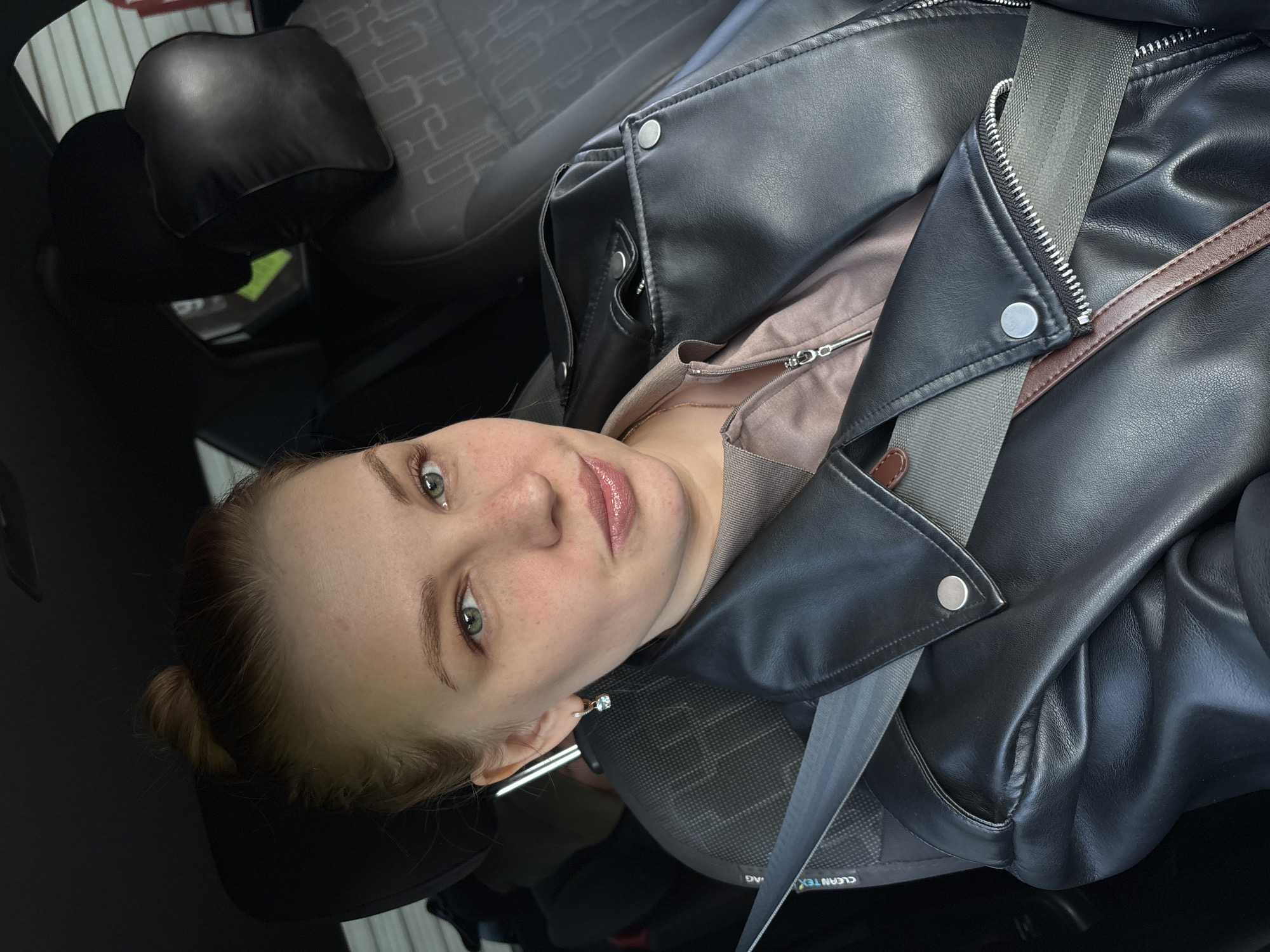 A young woman with fair skin and light brown hair pulled back into a bun is in the driver's seat of a car, wearing a black leather jacket over a light-colored top. A gray seatbelt is secured across her chest. The interior of the car is visible, including the gray patterned fabric of the seat and a black headrest. The image was taken inside a car, with the focus on the woman. The location and city, Rusanovo, Russia, are not discernible from the image alone.