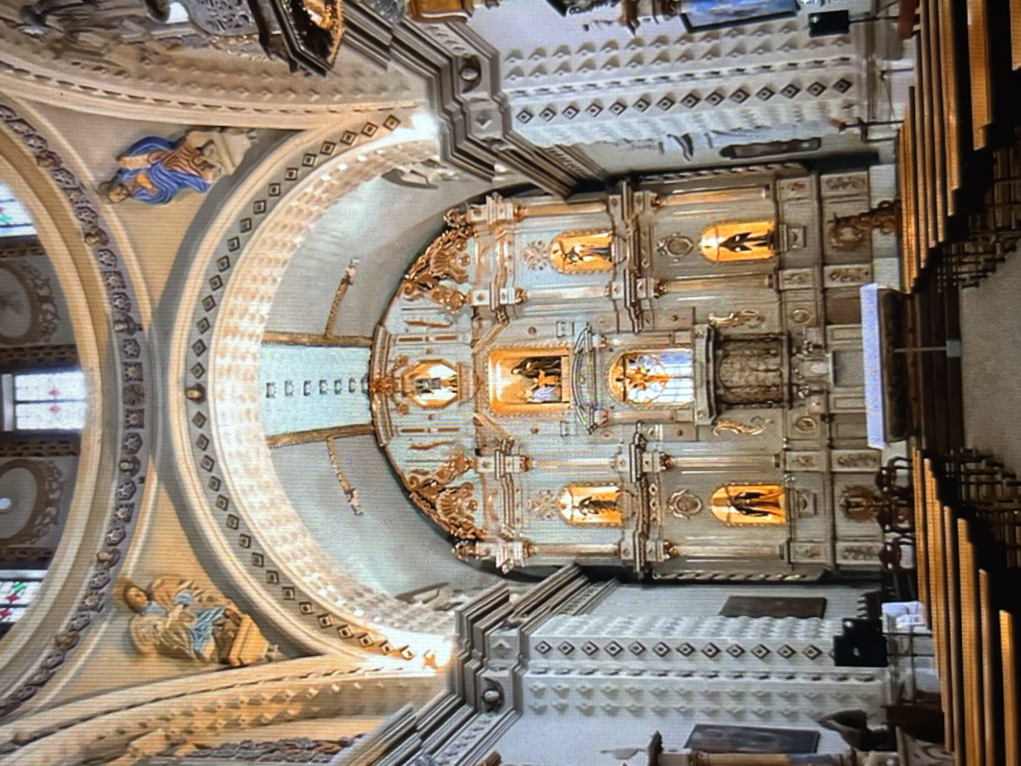 This is an interior shot of a church, likely taken looking upwards towards the altar area. The location is Tinaquillo, Venezuela. The building exhibits ornate architecture with arches, detailed moldings, decorative paintings or carvings of figures, and elaborate gold-colored accents. A large pipe organ is visible on the right side. The main altar has a multi-tiered design with framed artwork, possibly depicting religious scenes. The overall environment suggests a significant religious structure in Tinaquillo, Venezuela.