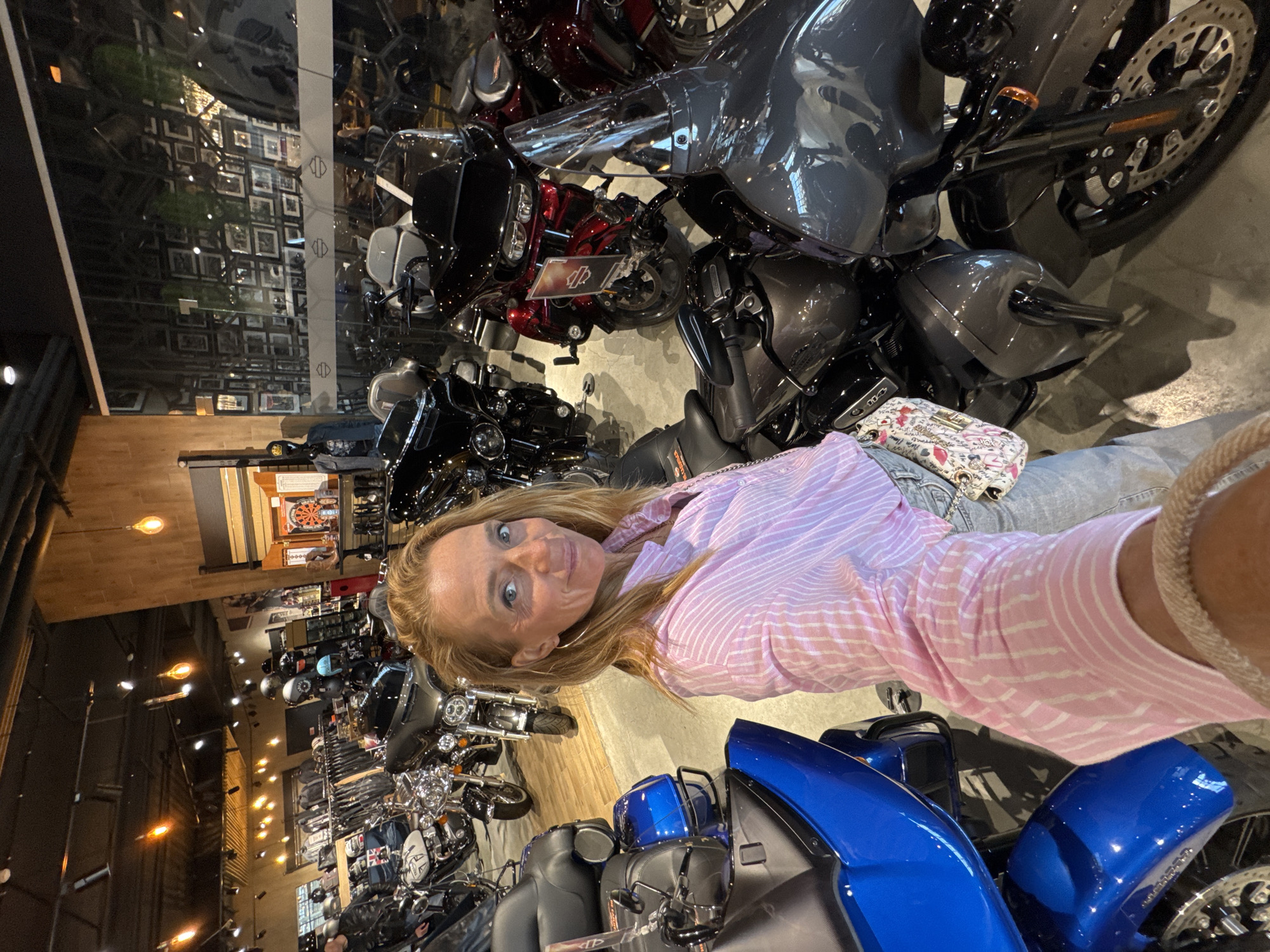 The image shows a fair-skinned woman with blonde hair standing inside a motorcycle dealership in Saint Petersburg, Russia. She is wearing a pink and white striped shirt, blue jeans, and a woven bracelet on her right wrist. A small purse is visible on her left side.
She is surrounded by several motorcycles, mainly Harley-Davidson models, in various colors including gray, black, red, and blue. The dealership appears to be well-lit with numerous display motorcycles and other merchandise.
The scene is reflective, with mirrors and glass surfaces showing reflections of the showroom and displays. In the background, framed pictures and decorations line the walls.