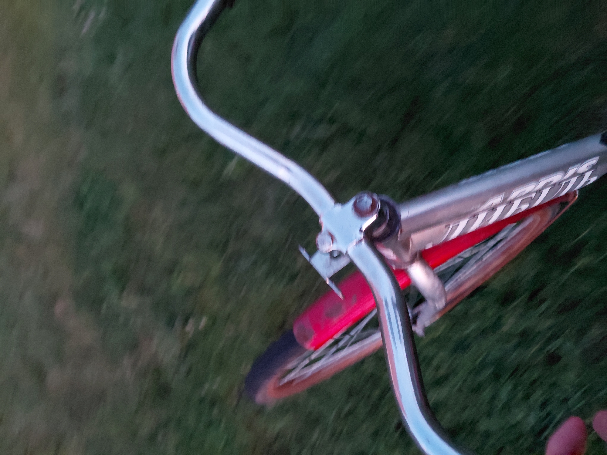 The image shows a first-person perspective of someone riding a bicycle. The handlebars are prominently featured in the foreground, with a chrome finish and a curved shape. The bicycle frame is visible below the handlebars and appears to be silver with some text. A red fender is attached to the rear wheel, which is blurred due to motion. The bicycle is moving over a grassy area. There is no specific indication of the location being Ustyvytsia, Ukraine. The scene is outdoors, with the grass suggesting a park, lawn, or field. The lighting suggests it may be dusk or dawn.