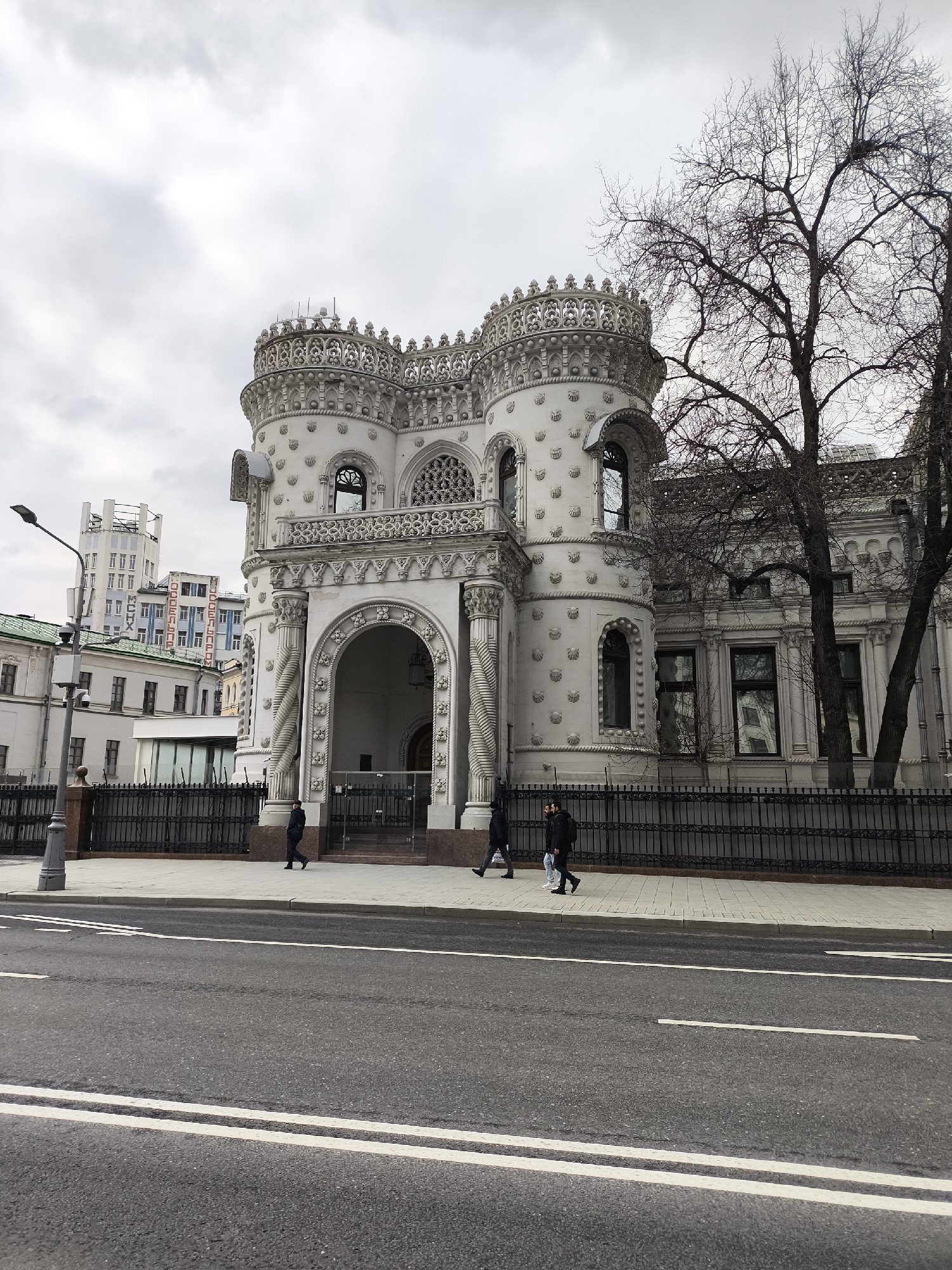 The image shows the Igumnov House, an ornate white building located in Moscow, Russia. It features distinctive turrets, intricate decorative elements, and arched entrances. A black wrought iron fence runs along the front of the building, separating it from the sidewalk.
Several pedestrians are walking on the sidewalk in front of the building. In the foreground is a wide paved road with white lane markings.
In the background, other buildings are visible, including a taller, modern structure with lettering in Cyrillic. The sky is overcast. A bare tree stands to the right of the Igumnov House.