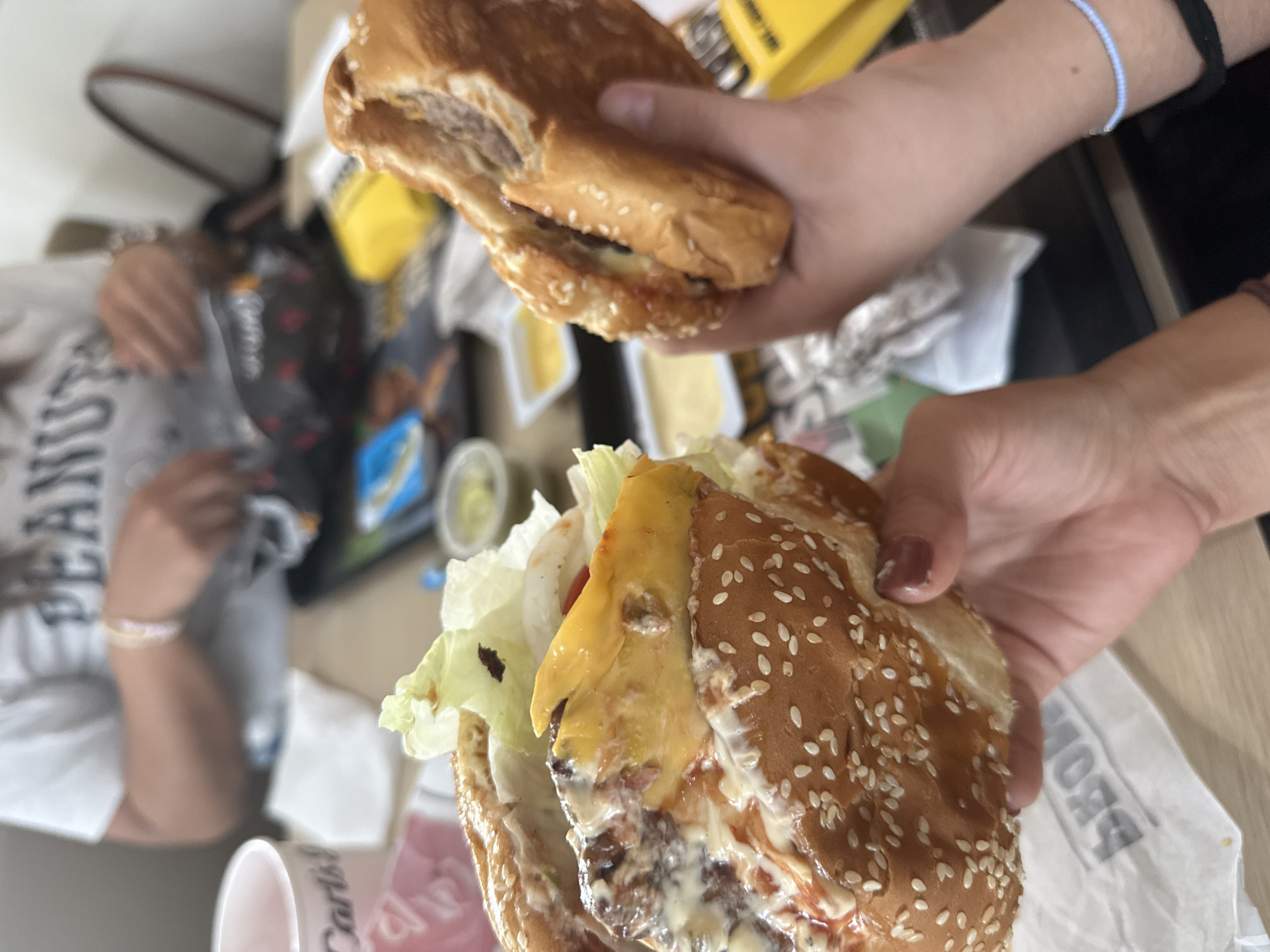 The image shows two people holding burgers, likely from Carl's Jr., given the branding on a nearby cup. One burger is closer to the camera, displaying lettuce, cheese, and sauce. The other burger is held higher, with a clear view of its sesame seed bun. The setting appears to be a fast-food restaurant, possibly a Carl's Jr. branch, considering the cup design and food items. The exact location is presumed to be San Bernardino Tlaxcalancingo, Mexico, based on the given data. Further detail about the environment is limited by the focus on the burgers and the blurred background. There are no apparent actions besides the holding of burgers. The interactions are limited to the individuals presumably eating.