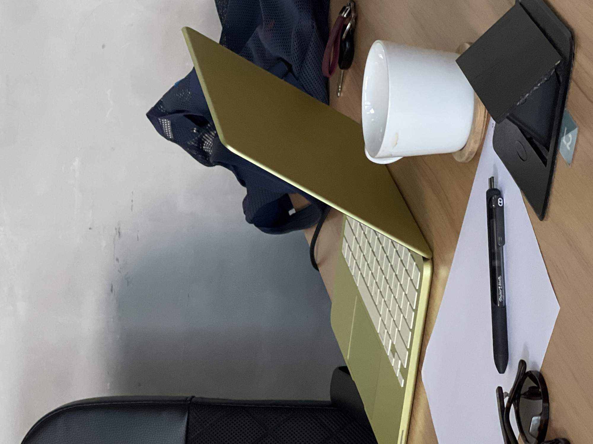 A close-up shot of a desk in General Luna, Philippines, reveals a workspace setup. A light green laptop is open, its keyboard and screen angled towards the viewer. To the right, a white mug with coffee stains sits on a wooden coaster. Nearby, a set of keys with a red keychain and a black car key are visible, along with a small black object that appears to be a phone stand or wallet. In the foreground, a white sheet of paper is laid out, with a black pen resting on top. A pair of dark-rimmed glasses is also present on the paper. In the background, a dark blue mesh bag and a portion of a black office chair are visible. The wall behind the desk has a textured, light gray finish with some faint marks.