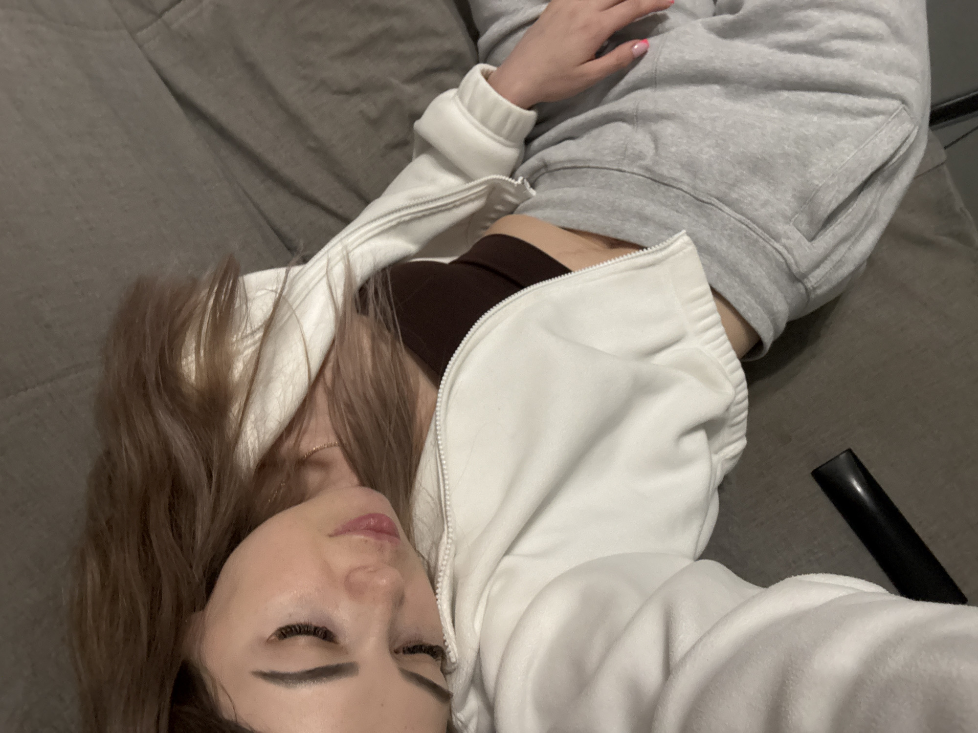 A woman is lying on a grey surface, likely a bed or couch, wearing a white zippered hoodie over a brown top, and grey sweatpants. She is positioned at an angle in the frame, with her head towards the lower left corner and her body extending diagonally. Her eyes are closed, and she appears to be resting. A black, cylindrical object is visible near her right side. The location is Unknown, Unknown.