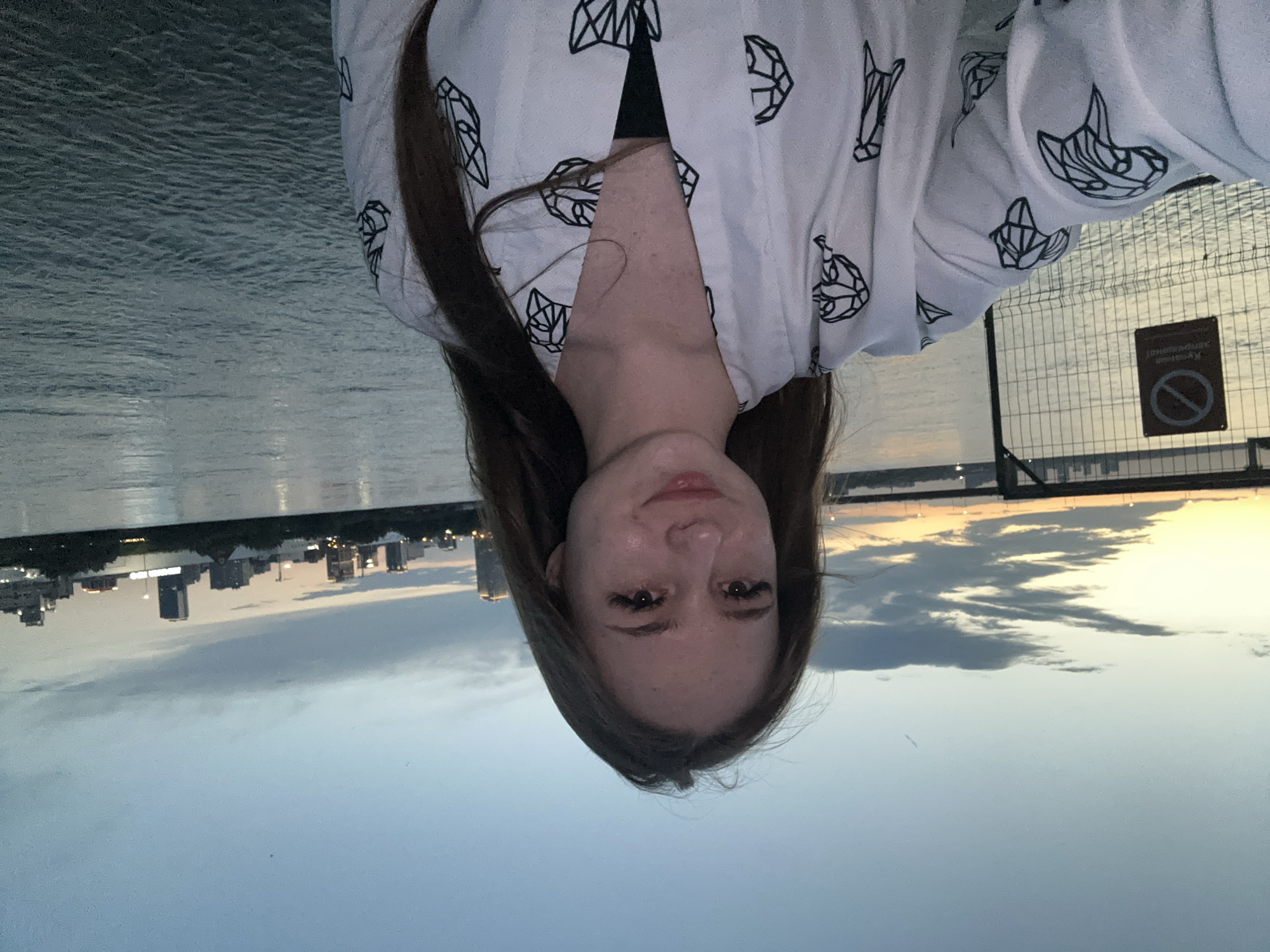 In the foreground, a young woman with long, brown hair and light skin takes a selfie outdoors at dusk. She is wearing a white robe with a geometric black pattern and a black top underneath. Her eyes are closed, and she is smiling faintly.
Behind her, a large body of water reflects the dim evening sky. In the distance, a city skyline with several buildings is visible, with lights twinkling from the structures. A bridge spans the water in the mid-ground, and to the left, a fence with a "no entry" sign is partially visible. The sky is a gradient of blue and orange, indicating the sunset.
