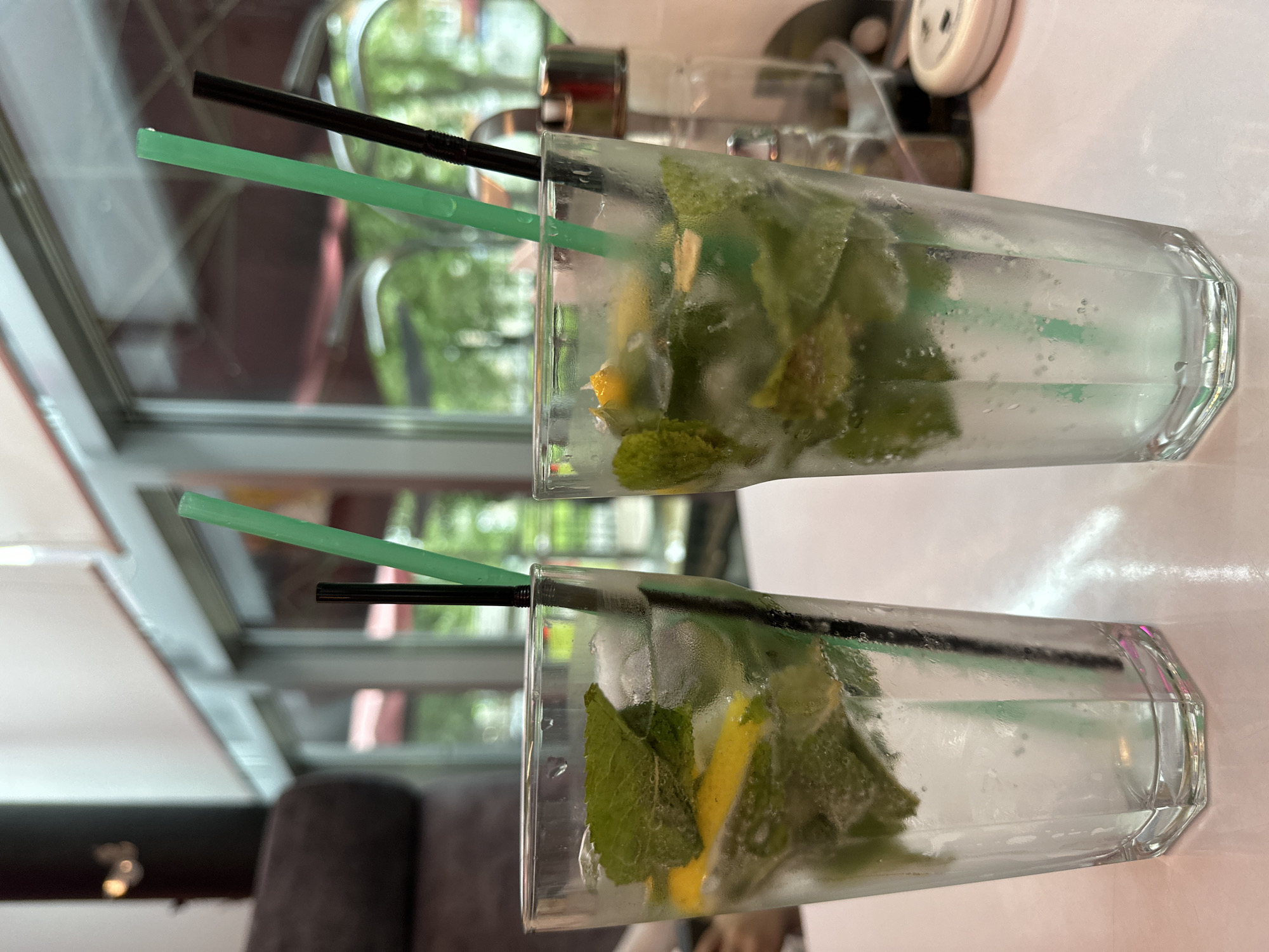 Two tall glasses of mojitos are displayed, each containing mint leaves and lemon slices. One glass is positioned in the foreground, slightly to the right, and the other is behind it and to the left. Both glasses have condensation on the outside, indicating they are cold. Each drink has two straws: one black and one green. The background appears to be a restaurant or cafe setting, with reflections in the window showing greenery and outdoor seating, suggesting an outdoor seating area or a view of trees. The lighting suggests it is daytime. There is no visible text in the image. The scene is calm and suggests a relaxing moment enjoying beverages.