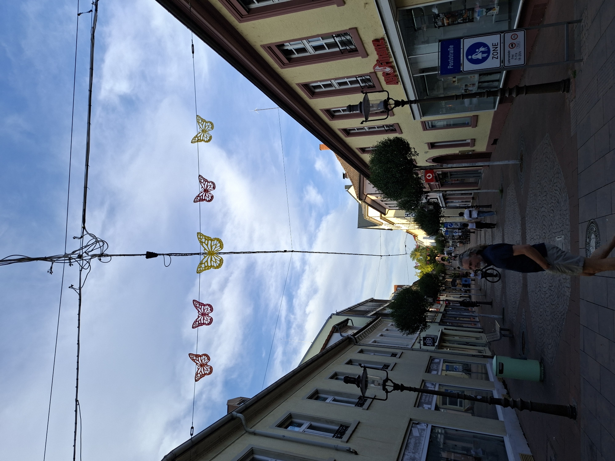 The image depicts a street scene in Rastatt, Germany, likely in the city center, given the commercial establishments and pedestrian activity. Overhead, a network of wires crisscrosses the sky, some with visible junctions and insulators. Strung between the wires are decorative, colorful paper butterflies, predominantly yellow and red. The street itself is paved with cobblestones and lined with buildings on both sides.

On the left, a pale yellow building features multiple windows and a prominent lamppost with a classic design. Shops with large glass windows are visible, one with text that appears to include "Digitalisieren." Further down, a light green trash receptacle stands near a bench.

On the right, a yellow building with red trim displays various shop fronts. One prominent sign reads "ABC Finanz." Further along, a blue and white sign indicates a "Poststraße ZONE" with symbols for pedestrian and child access, and a separate sign indicating delivery vehicle restrictions from 7 AM to 7 PM, with a weight limit of 7.5 tons.

Several people are present. In the foreground, a man with long, gray hair and a beard walks towards the right, wearing a dark blue polo shirt and light-colored shorts. He has his hands in his pockets. Further down the street, other individuals are seen walking, and a person is riding a bicycle. The sky is partly cloudy with a bright blue expanse. The overall impression is a somewhat quaint European street, possibly a shopping area, decorated for an event or season.