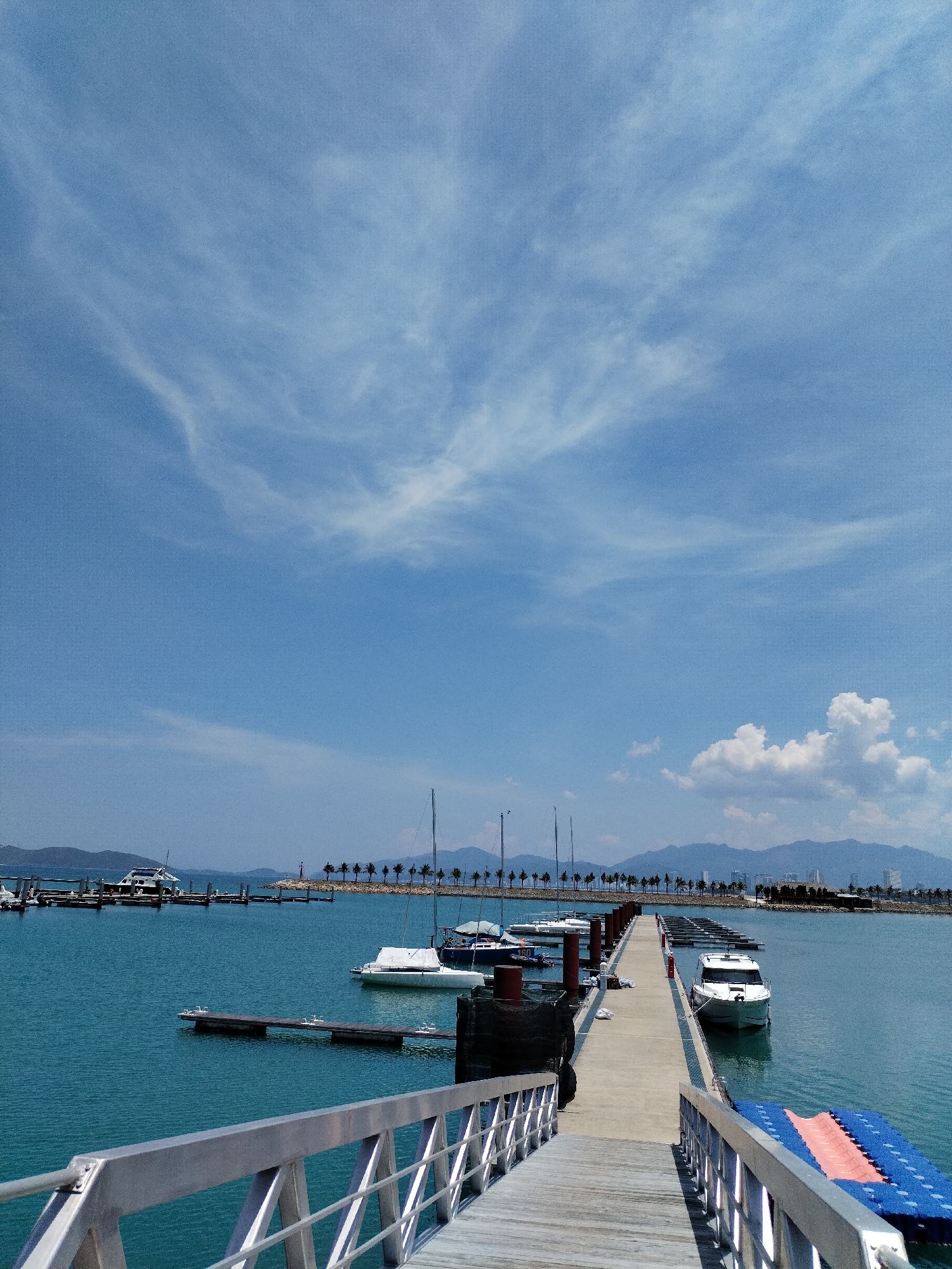 The image depicts a marina in Nha Trang Ward, Vietnam. A concrete pier extends into the turquoise water, flanked by docked boats. The pier is accessed by a wooden ramp with aluminum railings. A variety of boats are moored, including sailboats and motorboats. In the background, a coastal skyline is visible, featuring mountains and buildings. Palm trees line the distant shore. The sky is mostly blue with scattered cirrus clouds and cumulus clouds.