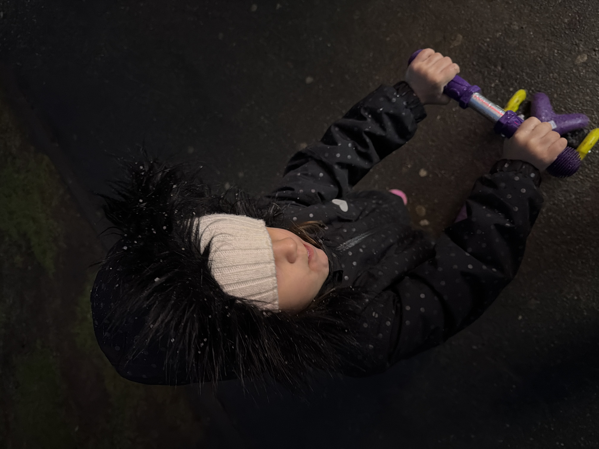 An overhead shot captures a young child holding the purple handlebars of a scooter outdoors, likely in the evening or at night. The child is wearing a dark, polka-dotted winter coat with a fur-lined hood and a light-colored knitted beanie. Small white specks, consistent with snowflakes or sleet, are visible on her hat, fur, and coat, suggesting cold or wet weather. Her face is partially visible, showing a calm expression.
The child's hands firmly grip the purple handlebars of the scooter, which feature yellow rubber grips at the ends and a central metallic section. The background is a dark, possibly wet, paved surface, with a dimly lit patch of green vegetation visible on the left. The overall lighting is dim, indicating it's dark outside. No legible text is visible on the scooter or elsewhere in the image. The scene suggests a momentary pause during outdoor play in wintery conditions.