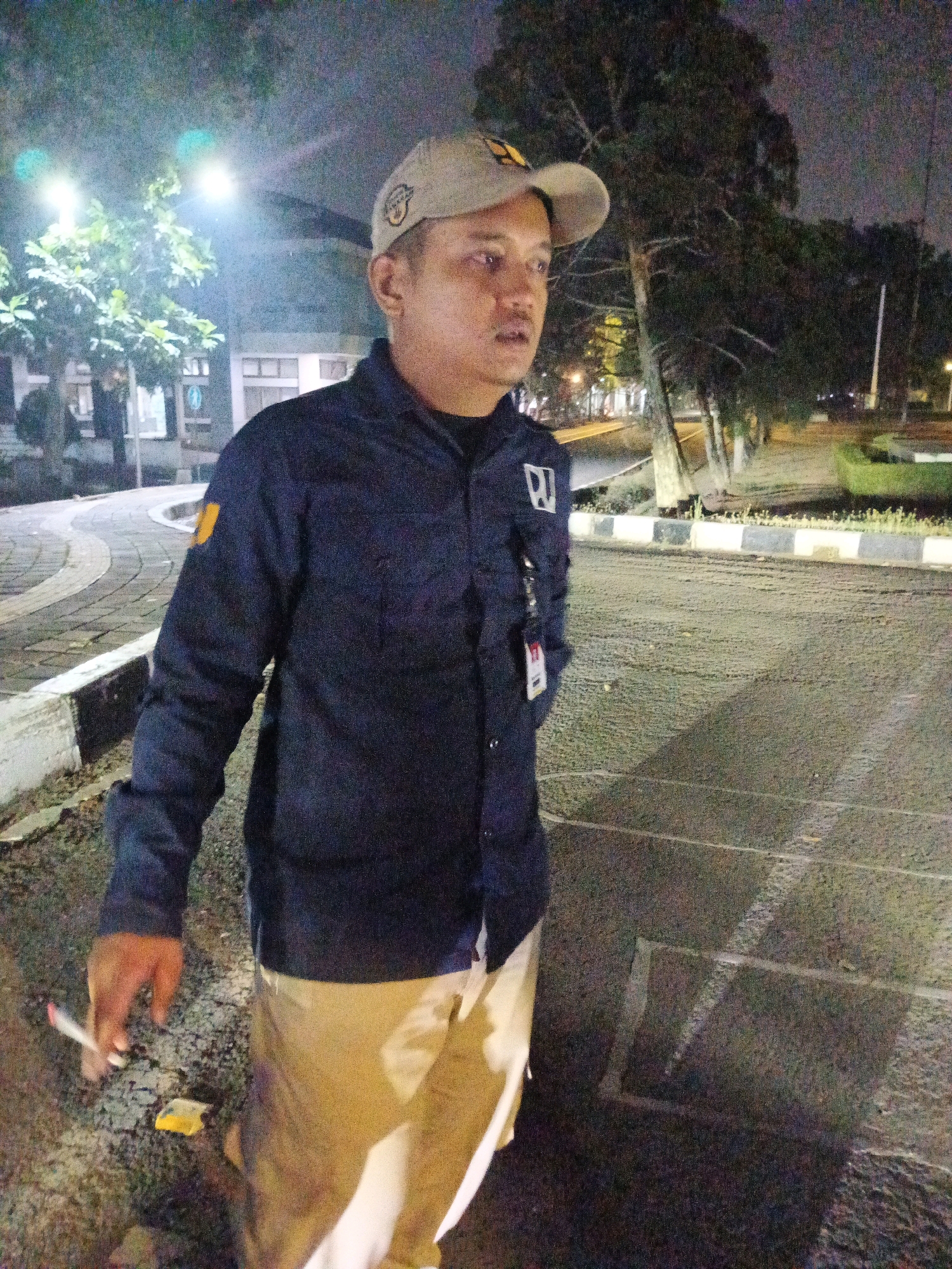 The image shows a man standing outdoors at night. He is wearing a dark blue long-sleeved shirt with a yellow emblem on the left shoulder and another emblem on the left chest. He also wears a light-colored baseball cap and light-colored pants. The man is holding a cigarette in his left hand, which is lit. In the background, there are trees, streetlights, and buildings. The ground appears to be asphalt or concrete with some paving stones visible to the left. There is no specific indication of the location being within Bandung City, Indonesia.