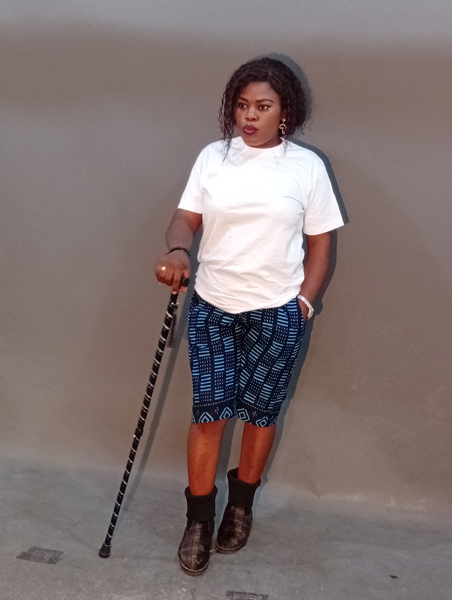 A full-body, medium shot captures a Black woman standing against a plain, grey backdrop. She has curly, dark hair and is wearing a plain white t-shirt, blue patterned shorts, and dark, fur-lined boots. The woman is holding a black and silver patterned walking cane in her left hand, with her right hand in her pocket. Her left arm rests on the cane, and her right hand is out of view in her pocket. She is looking towards her right, with her mouth slightly open. The environment appears to be an indoor studio setting with a concrete floor. The lighting is even, creating soft shadows. The scene is static and posed, suggesting a portrait or fashion photograph.