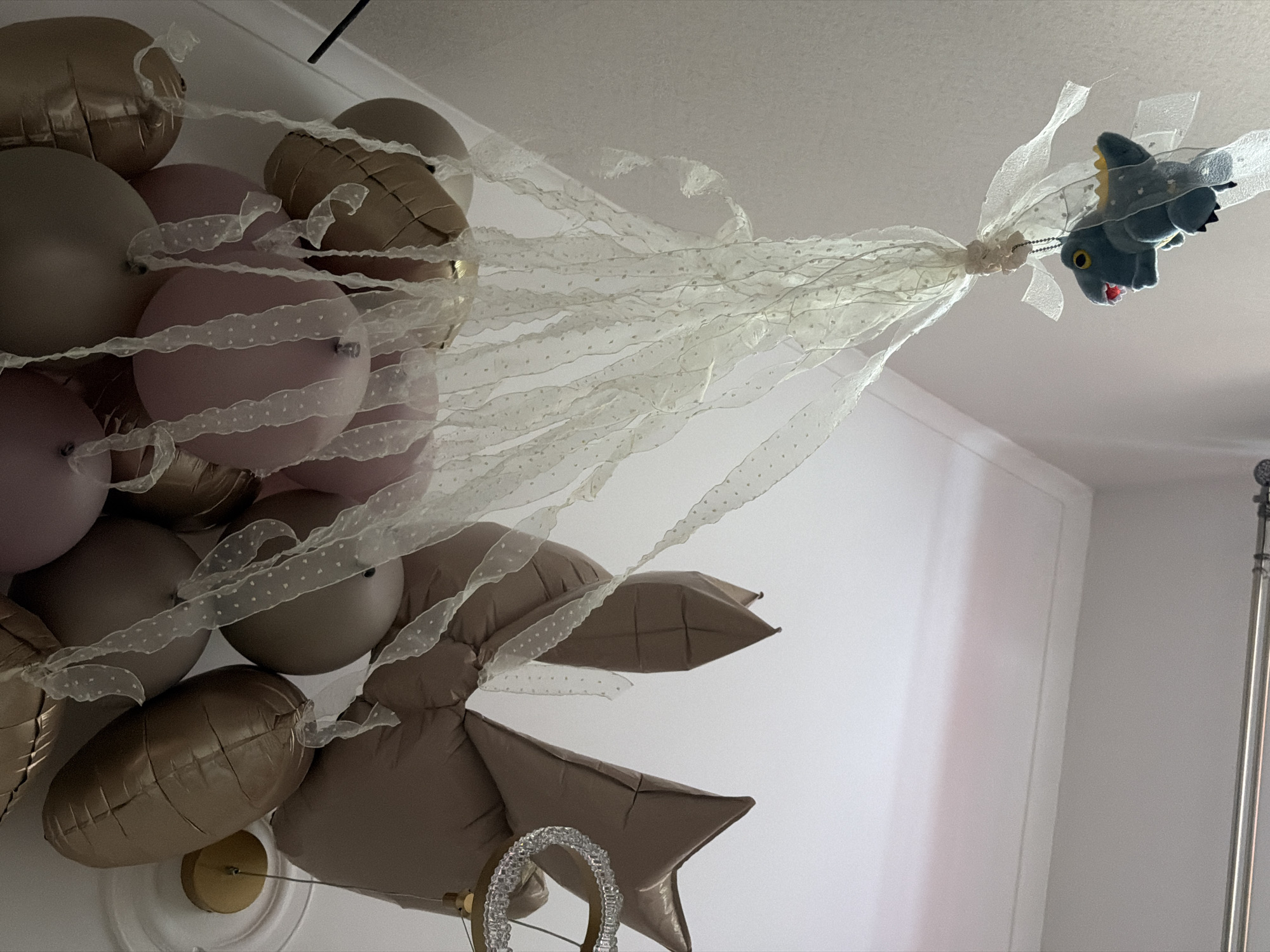The image shows a cluster of balloons in shades of pink, brown, and gold attached to a small gray stuffed animal, possibly a dragon or dinosaur. The balloons and stuffed animal are connected by white ribbon with small dots. The scene appears to be indoors, possibly a decorated room, with parts of a white wall and a silver pole visible in the background. The image's composition suggests the scene is viewed from below, looking upward towards the ceiling where the balloons are likely attached. There is no indication of the location being Moscow, Russia, or any specific city. There are no people in the image.