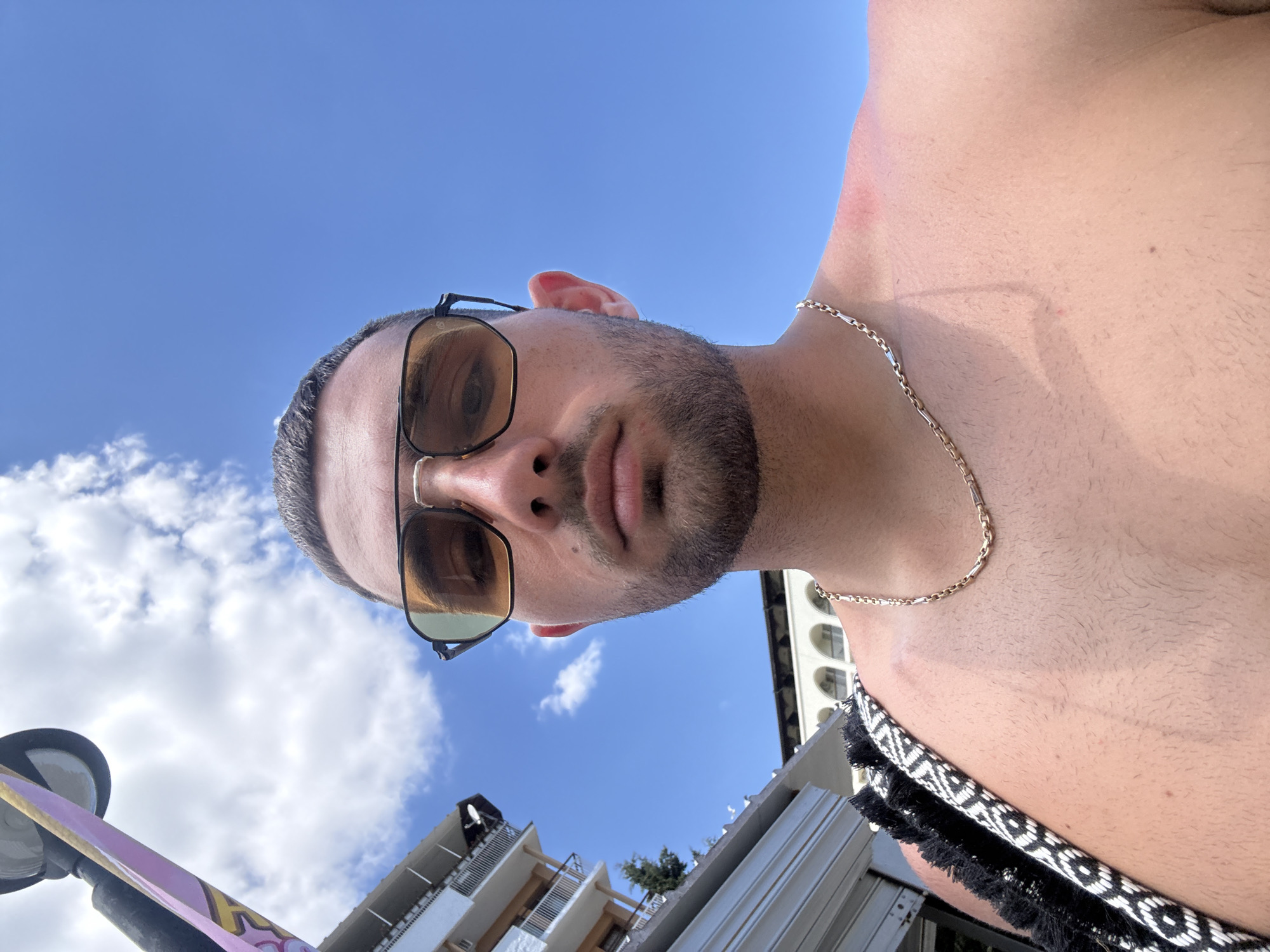 The image is a close-up, low-angle selfie of a man with a beard and sunglasses, looking directly at the camera. He is shirtless, wearing a gold chain, and has a bag with a black and white patterned strap over his shoulder. The background shows a bright blue sky with scattered clouds, and the upper levels of buildings, suggesting an outdoor urban or resort setting. The lighting indicates it is daytime. Based on the architectural style and the known landscape of Nesebar, Bulgaria, the buildings in the background are likely part of the historical architecture found in the old town, possibly overlooking the sea. The man's skin appears tan, and there are visible tan lines on his shoulder and chest.
