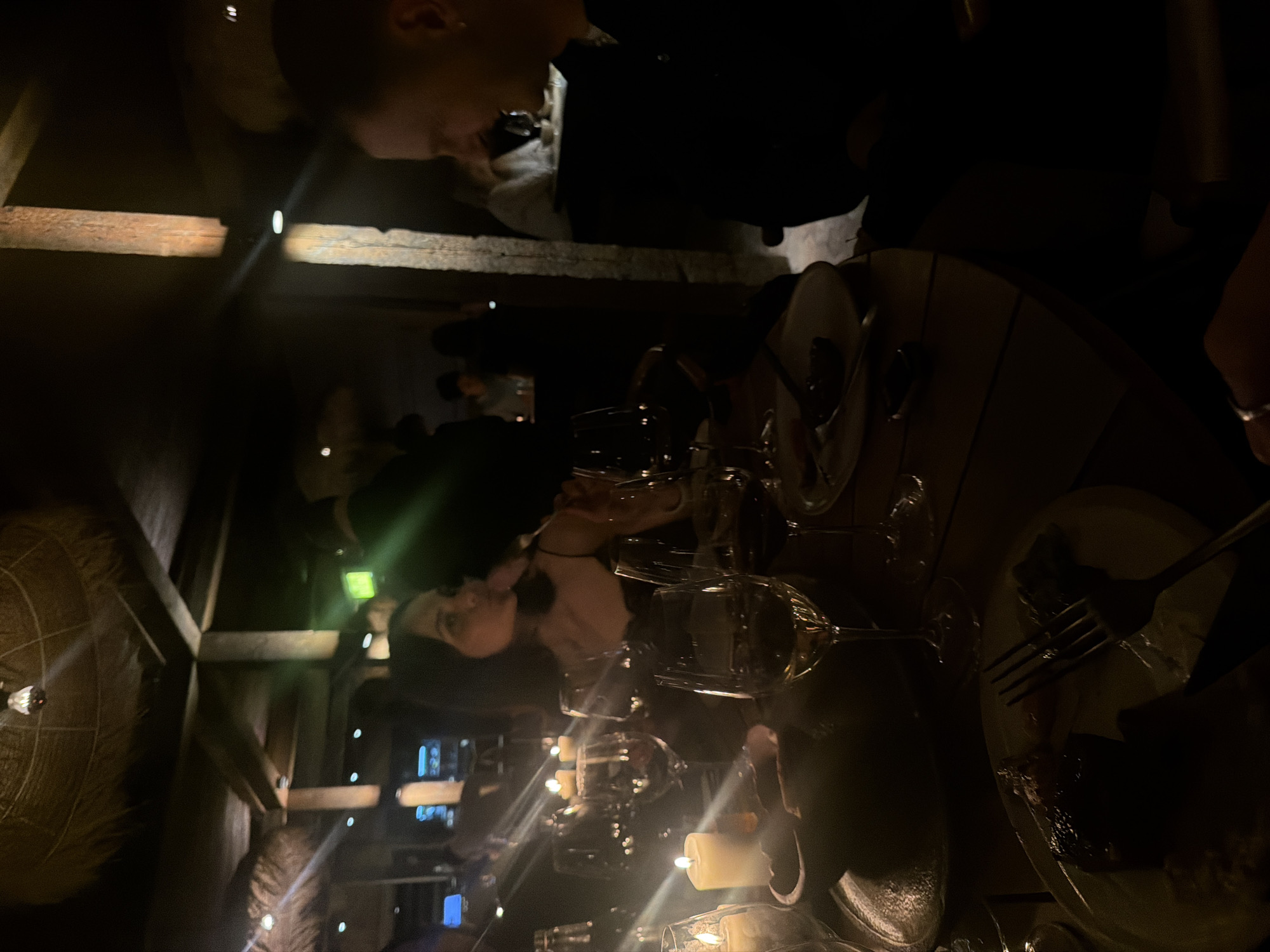 The scene depicts a dimly lit restaurant environment, possibly in London, United Kingdom. The lighting is low, creating a dark ambiance. Several individuals are seated around a wooden table, with visible plates of food and cutlery suggesting a meal in progress. Wine glasses are arranged, some illuminated by candlelight. The restaurant has rustic decor with wooden beams and what appears to be woven details on the walls. The people seem to be engaging in conversation while dining. There's a notable glow and flare from the light sources, indicative of low light levels. Other restaurant patrons can be seen in the background.