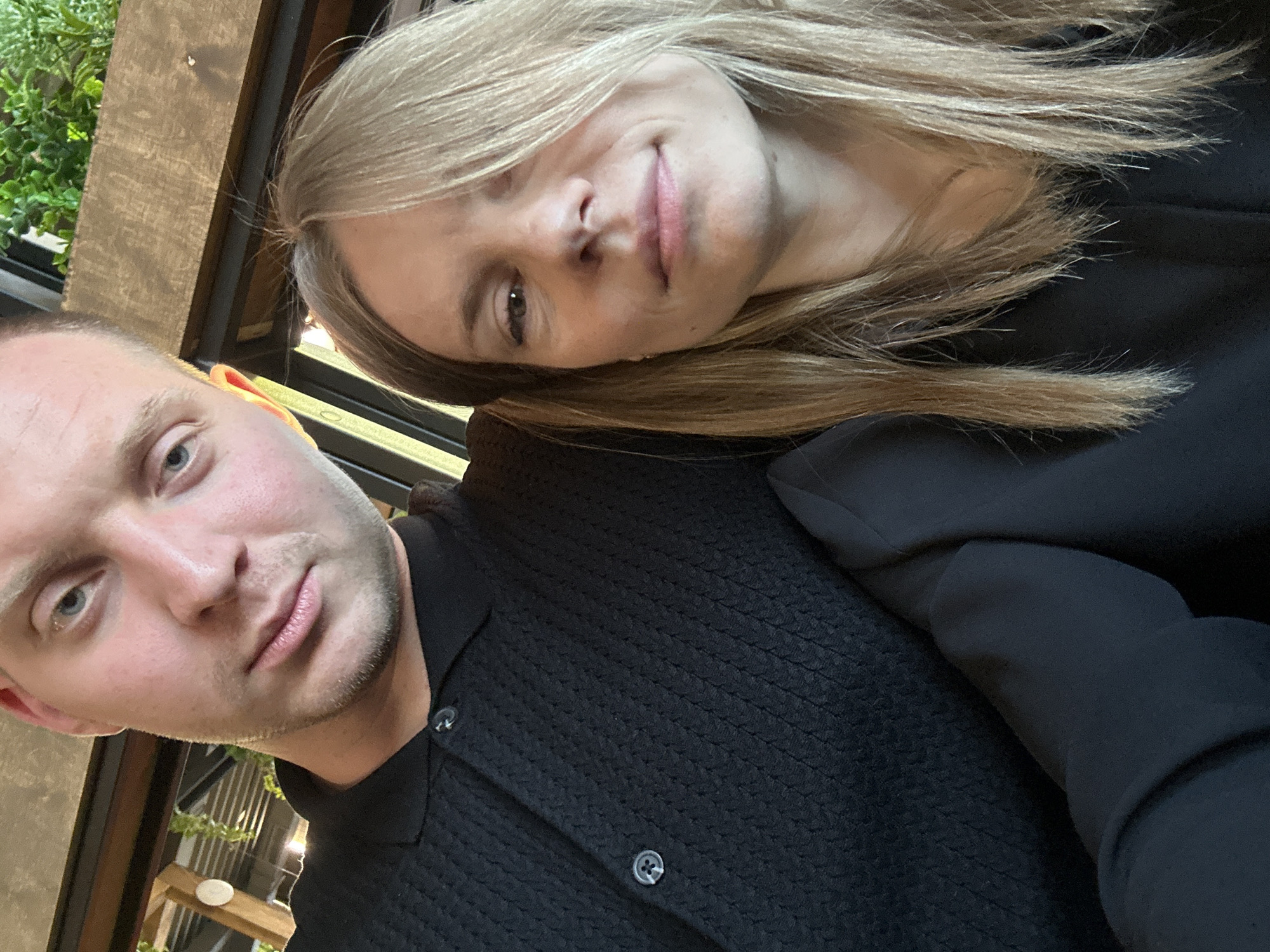 A man and a woman are taking a selfie. The man, on the left, is wearing a black polo shirt with a textured pattern and two buttons at the neck. He has fair skin, light blue eyes, and short, light brown hair. He is looking directly at the camera. The woman, on the right, has fair skin, light brown hair that falls over her face, and is looking slightly to the left. She is wearing a black top. The background appears to be an indoor setting, possibly a restaurant or cafe, with wooden beams and green plants visible. The image was taken on an overcast day. The city of Blagoveshchensk, Russia is not discernable from the image.