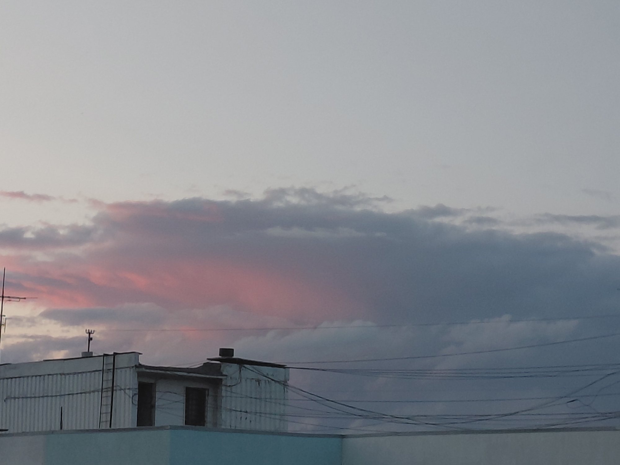 The image captures an urban sky at dusk, with a vast expanse of pale gray sky dominating the upper portion. A band of clouds stretches across the middle of the frame, transitioning from a soft pink hue on the left to darker grays on the right. The lower portion of the image features the rooftops of several structures, including buildings with corrugated metal siding and flat roofs. Wires are strung between poles, crisscrossing the sky above the buildings. An antenna-like structure is visible on one of the rooftops. The overall scene suggests an ordinary evening in an urban environment.