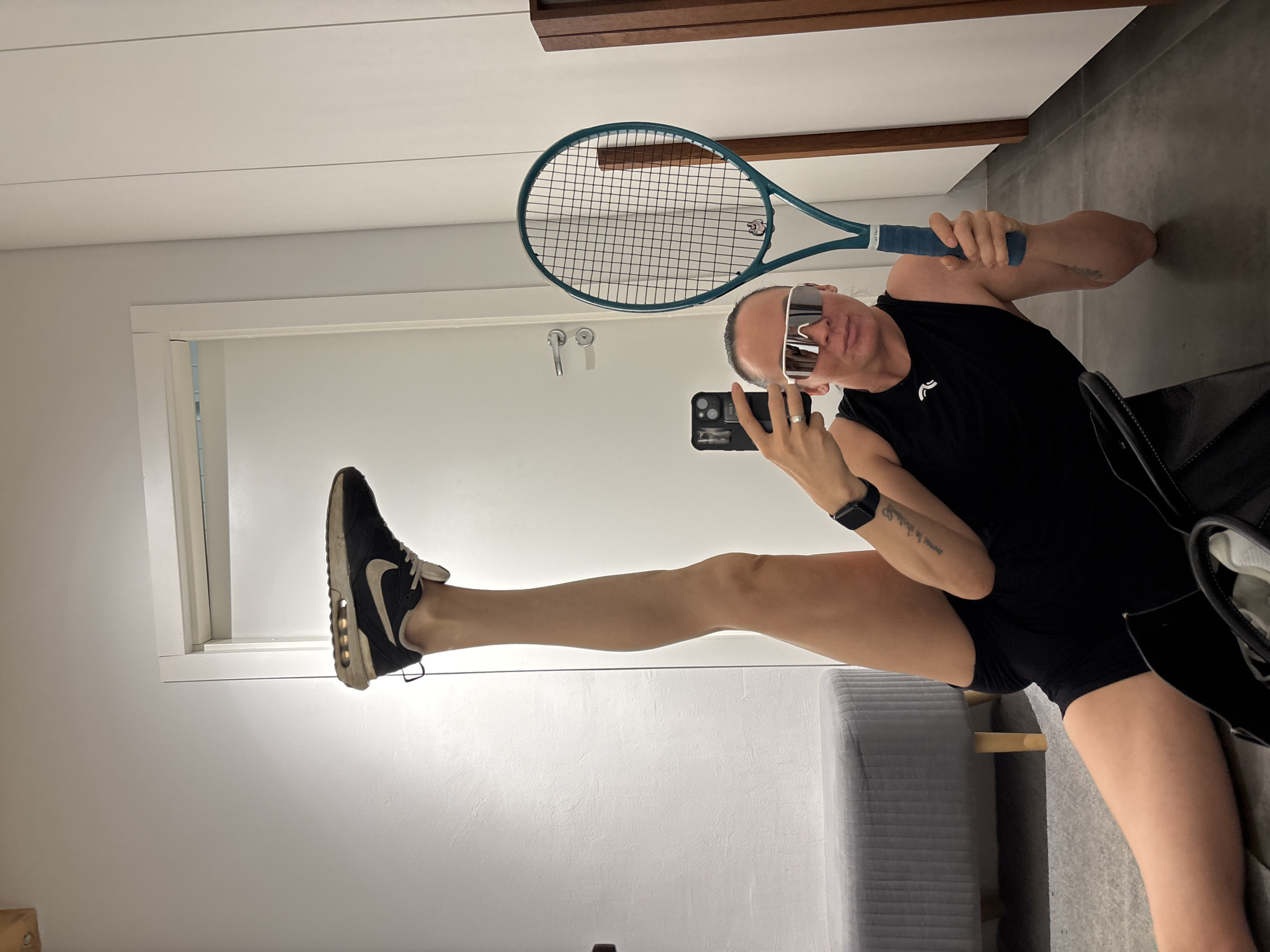 A person is taking a selfie in a room that appears to be inside a house or apartment. The person is wearing a black tank top, black shorts, sunglasses, and a watch. They are holding a smartphone in one hand and a teal tennis racket in the other. One leg is extended upwards towards the door frame, with a black Nike sneaker on the foot. The room contains a white door, grey walls, grey flooring, a grey bench, and a wooden beam near the ceiling. The exact location within the city of Chaikovsky, Russia cannot be determined from the image.