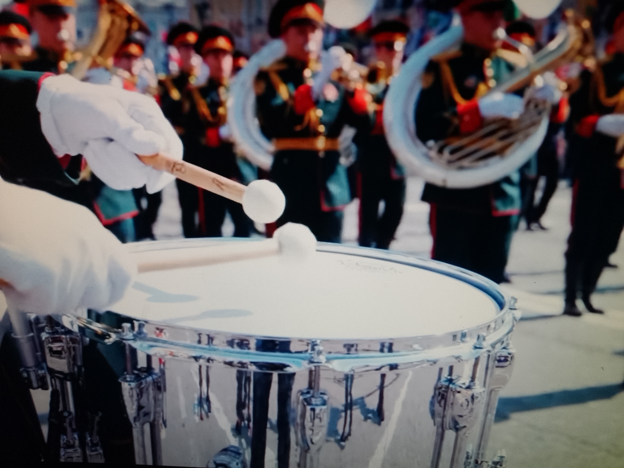 The image shows a close-up of a snare drum being played by a person wearing a white glove. The drum is silver and has chrome hardware. In the background, a marching band is visible, with members in uniform playing brass instruments and drums. The band members are wearing dark green uniforms with red accents and dark hats. The scene appears to be outdoors, possibly during a parade or public event. There is no visual information in the image that directly indicates the location as Chiajna, Romania.