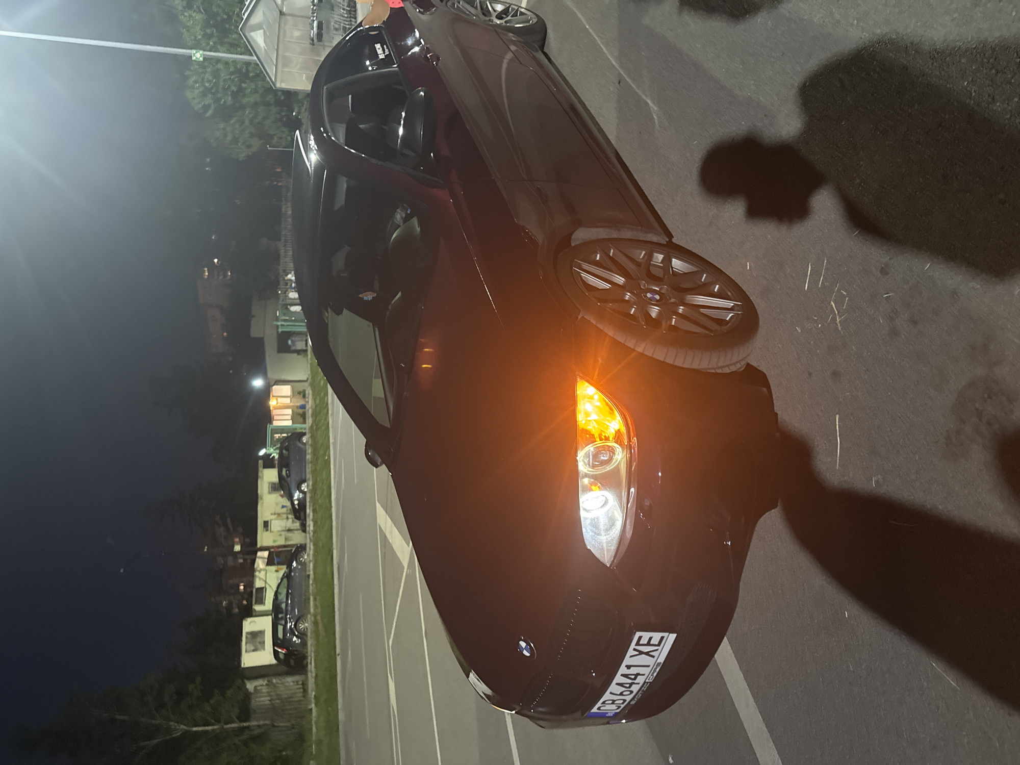 The image shows a dark-colored BMW convertible with its front right turn signal illuminated. The car is parked in a parking lot at night. The license plate reads "CB 6441 XE". The city and state are unknown.