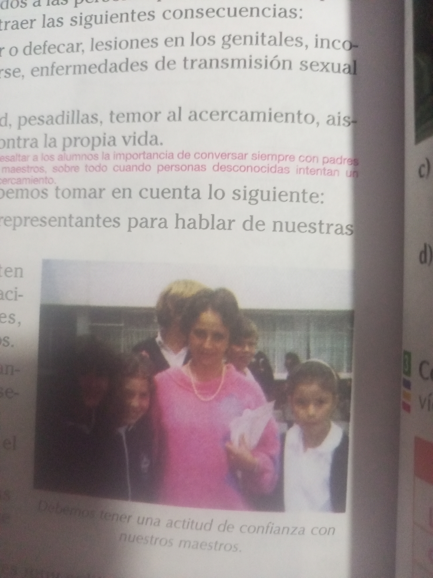 The image shows a page from a book. The text mentions "traer las siguientes consecuencias:", "r o defecar, lesiones en los genitales, inco-", "rse, enfermedades de transmisión sexual", "d, pesadillas, temor al acercamiento, ais-", "contra la propia vida.", "esaltar a los alumnos la importancia de conversar siempre con padres", "maestros, sobre todo cuando personas desconocidas intentan un", "cercamiento.", "bemos tomar en cuenta lo siguiente:", "representantes para hablar de nuestras", and "Debemos tener una actitud de confianza con nuestros maestros.". There are also two letters "c)" and "d)" and some text related to "Con Viá".
In the center of the page, there is a photograph of a group of people. In the foreground, a woman in a pink sweater is smiling and holding papers. To her left, a young girl in a dark jacket and white shirt is visible, and to her right, another young girl in a white shirt and dark jacket is looking at the camera. In the background, there are other people and a building.
The text on the page is in Spanish and discusses consequences, fears, and attitudes towards teachers and unknown individuals. The reference to La Tagua, Venezuela, is not present in the provided image.