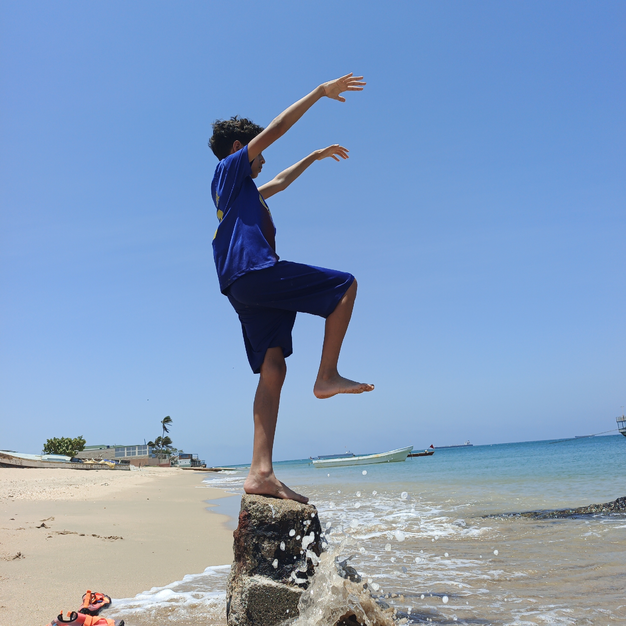 The image shows a young boy standing on a rock at the shoreline. He is balancing on one foot while raising his arms and the other leg. He is wearing a blue shirt and blue shorts. The location appears to be a beach, with sand and water visible. In the background, there are buildings and boats in the water. The image was taken in Villa Marina, Venezuela. The waves from the ocean are splashing against the rock. An orange pair of shoes are on the sand near the rock.
