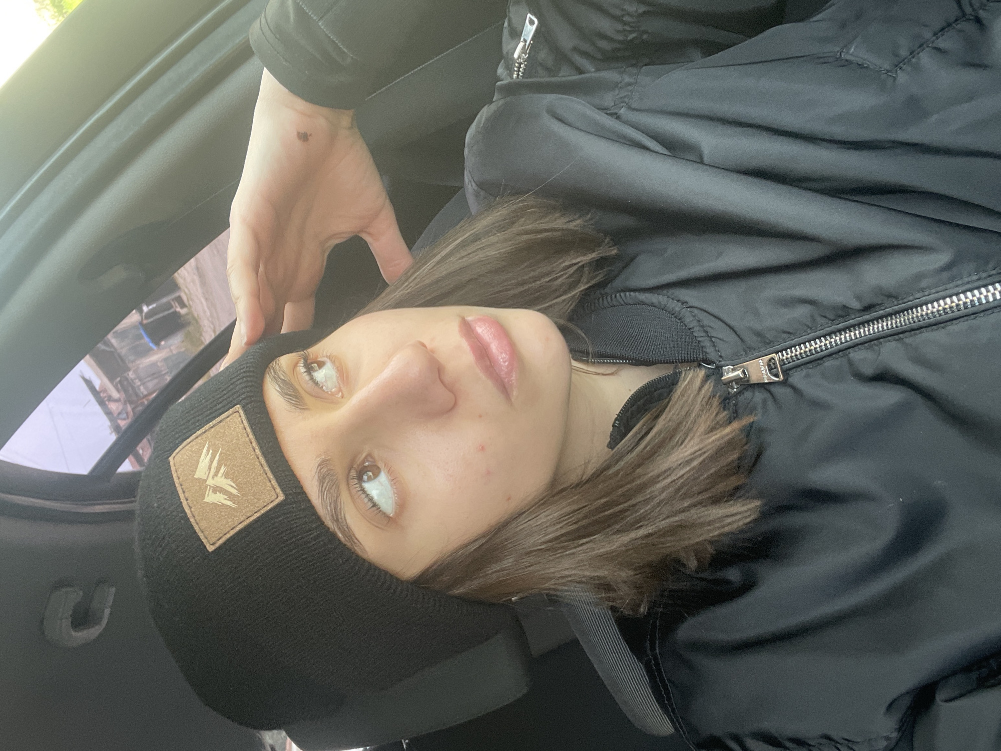 A young person, seen from an upward angle, is wearing a black beanie with a logo, and a black zip-up jacket. They appear to be inside a car, possibly in Sofia, Bulgaria. Their left hand rests on the interior of the vehicle. The exact location and the presence of Sofia, Bulgaria, are not identifiable from this image alone.