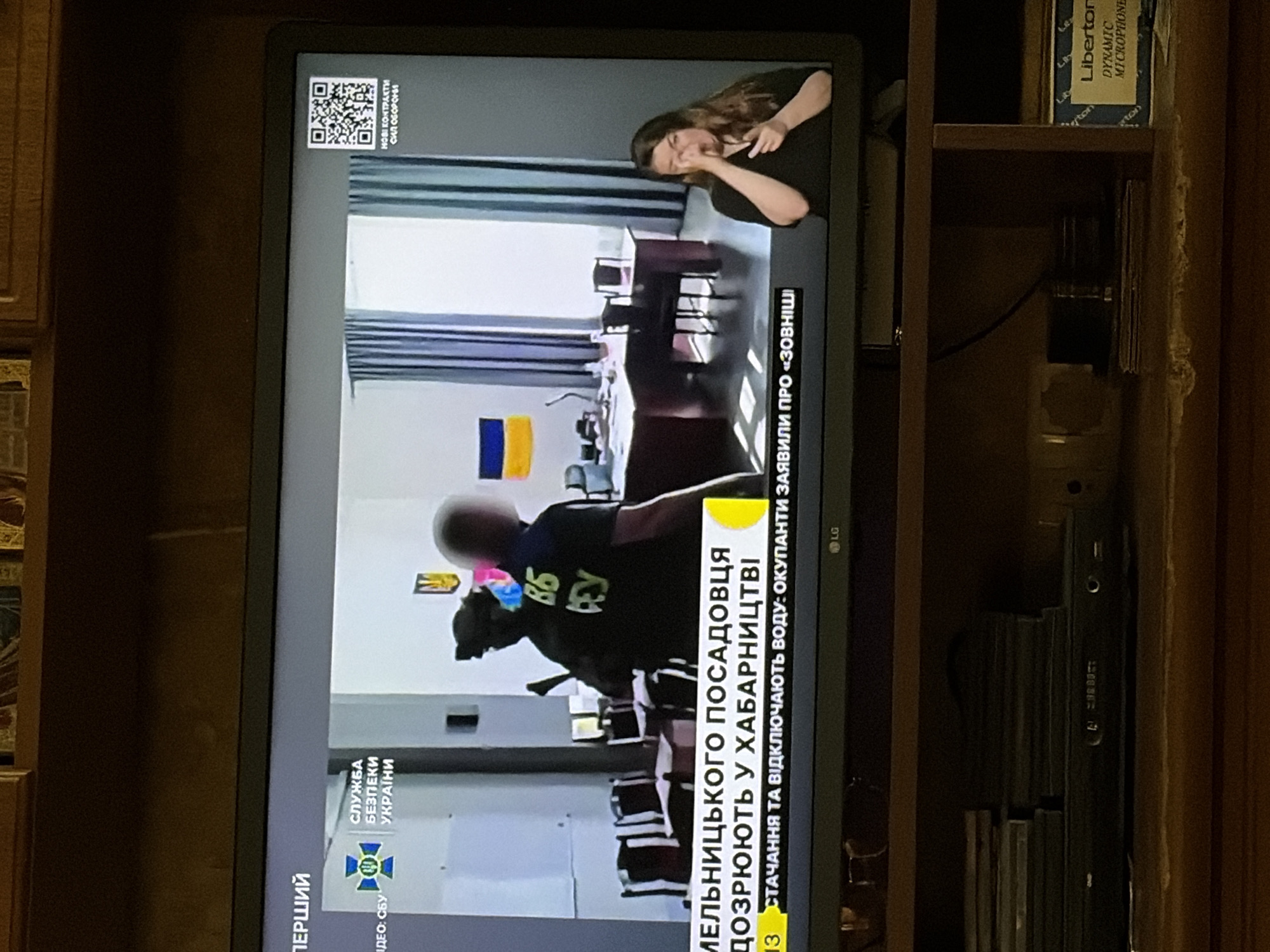 The image shows a television screen displaying a news broadcast. The screen shows a split view. On the left is a news report with Ukrainian text at the bottom. The text appears to be reporting on a case involving a local official and corruption. There is a logo for the "Service of Security of Ukraine" visible. On the right side of the screen, a woman is signing the news report in sign language. The background of the news report appears to be an office setting with desks, chairs, and curtains. The television itself is on a wooden shelf, and there are DVDs or CDs stacked below it. A box labeled "Liberton DYNAMIC MICROPHONE" is also visible on the shelf.