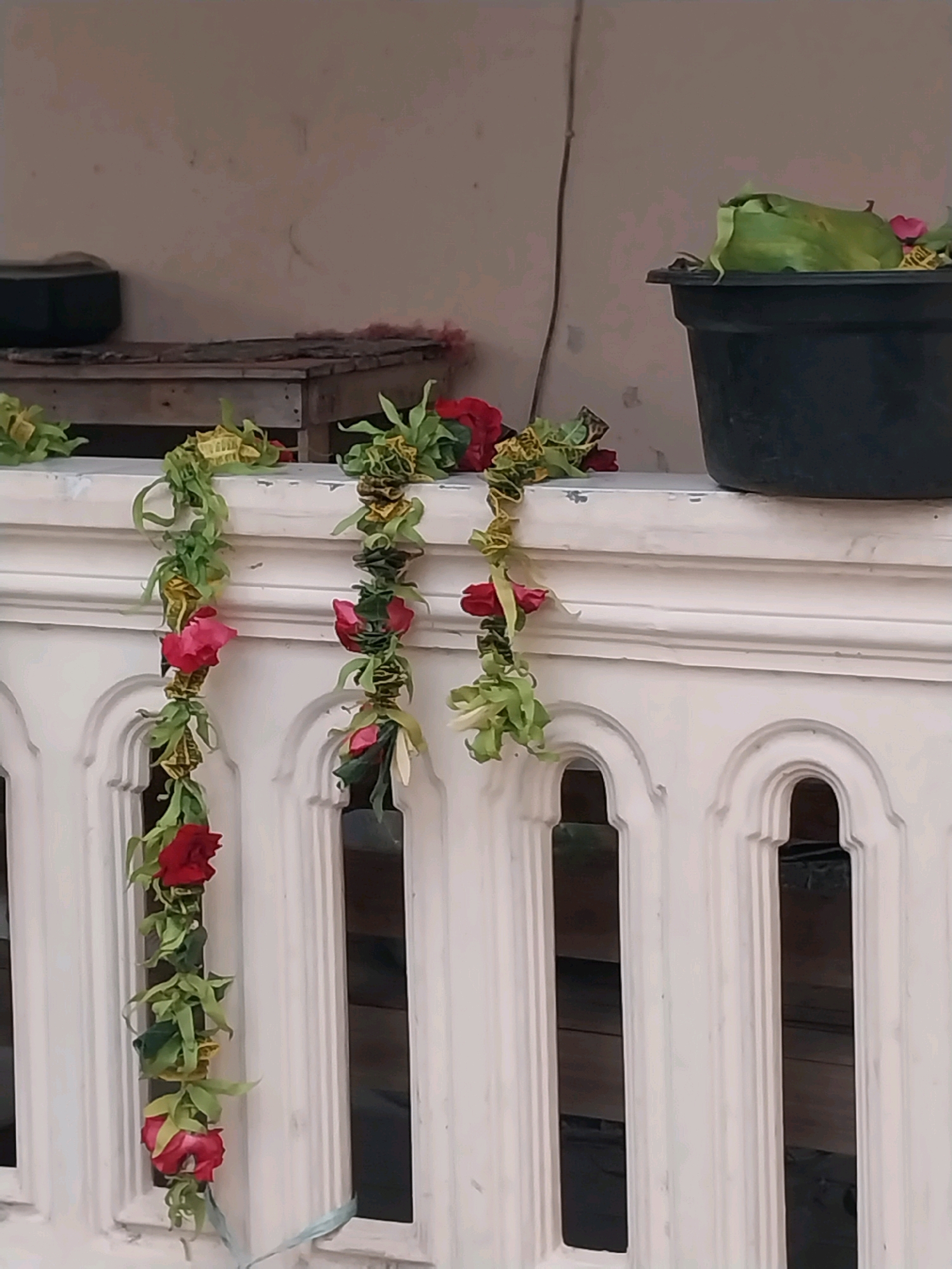 The image shows a decorative garland of artificial red roses and green leaves draped over a white railing. The garland hangs in vertical strands, with several roses visible along each strand. Behind the railing, a wooden structure and a black pot are partially visible. The scene appears to be outdoors, likely on a balcony or porch. The location is specified as Nglegok, Indonesia.