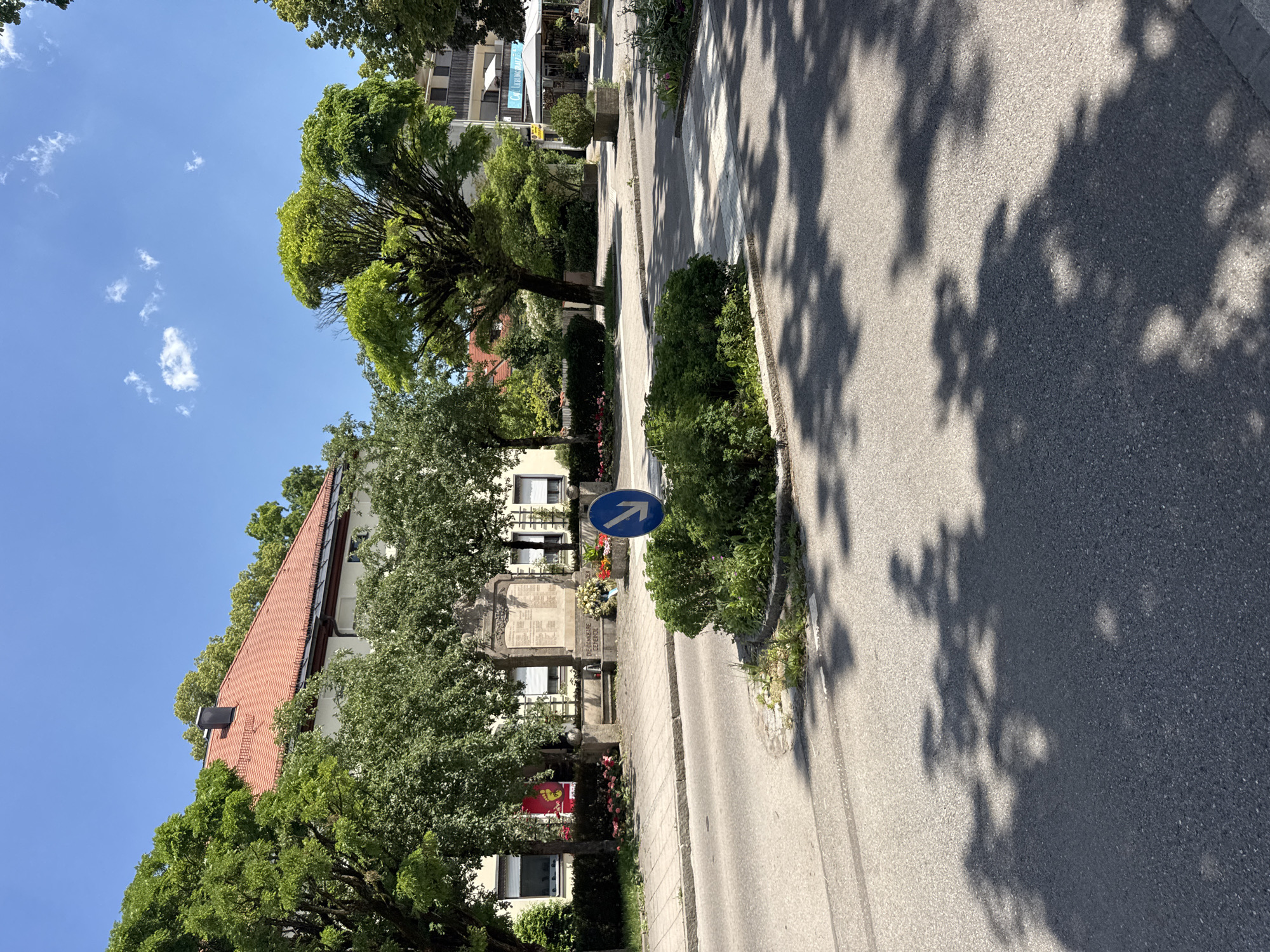 A vertical, eye-level shot captures a sunlit street scene in Höhenkirchen-Siegertsbrunn, Germany. The clear blue sky is dotted with wispy white clouds, suggesting a pleasant day. Lush green trees line the street, their leaves creating dappled shadows on the asphalt. In the foreground, a paved sidewalk runs alongside the road.
Dominating the left side of the frame is a building with a red-tiled roof, partially obscured by dense foliage. Further down the street, a war memorial stands prominently, adorned with flowers and what appears to be a plaque with names. A blue circular sign with a white arrow pointing upwards is mounted on a pole near the memorial. To the right of the memorial, a sidewalk with a pedestrian crossing is visible.
In the background, more buildings and trees can be seen, hinting at a residential or commercial area. The shadows cast by the trees suggest it is late morning or early afternoon. No people are visible in the image. The overall impression is one of a peaceful, sunny day in a German town.