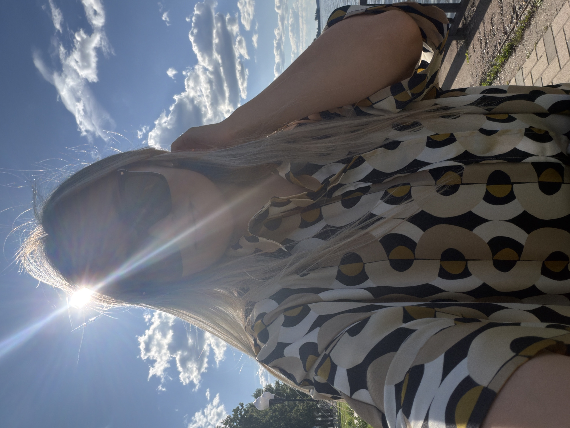 The image depicts an outdoor selfie taken by a person, likely female, with long blonde hair, wearing sunglasses, and a collared shirt with a geometric pattern in beige, black, white, and golden-yellow hues. The sun is positioned in the upper left, creating a prominent lens flare that obscures the face and upper body of the subject, indicating strong backlighting. The subject's right arm is visible, with the hand near the head, possibly adjusting hair or sunglasses.
The background shows a clear blue sky with scattered white cumulus clouds. To the left, green foliage and part of a white street lamp are visible, suggesting a park or landscaped area. To the right, a large body of water, possibly a lake or river, extends to the horizon, with indistinct land visible in the far distance. A dark metal railing or barrier runs along the edge of the water. The ground beneath the railing consists of a paved path (brick or cobblestone) and a strip of gravel or dirt with sparse green vegetation. The scene is located in the city of Votkinsk, Russia.