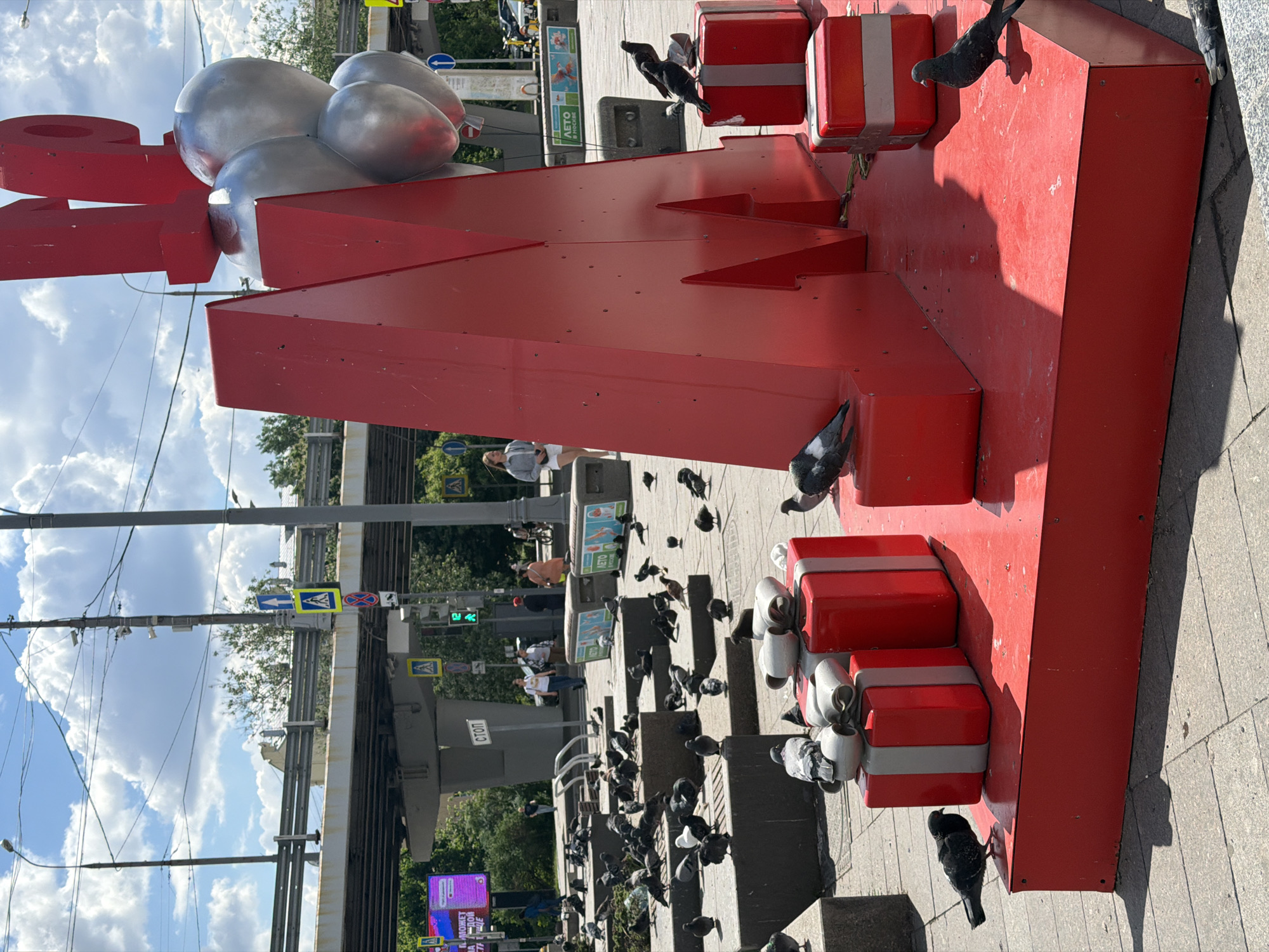 A daytime outdoor scene in Moscow, Russia features a large, red, "M" shaped sculpture decorated with silver "balloons" and several red rectangular shapes resembling gift boxes with gray bands. Numerous pigeons are scattered around the sculpture and the surrounding sidewalk. In the background, there is a pedestrian overpass, traffic lights, signage in Russian, and greenery. Several people are walking on the overpass and sidewalk. The sky is visible and has scattered clouds.