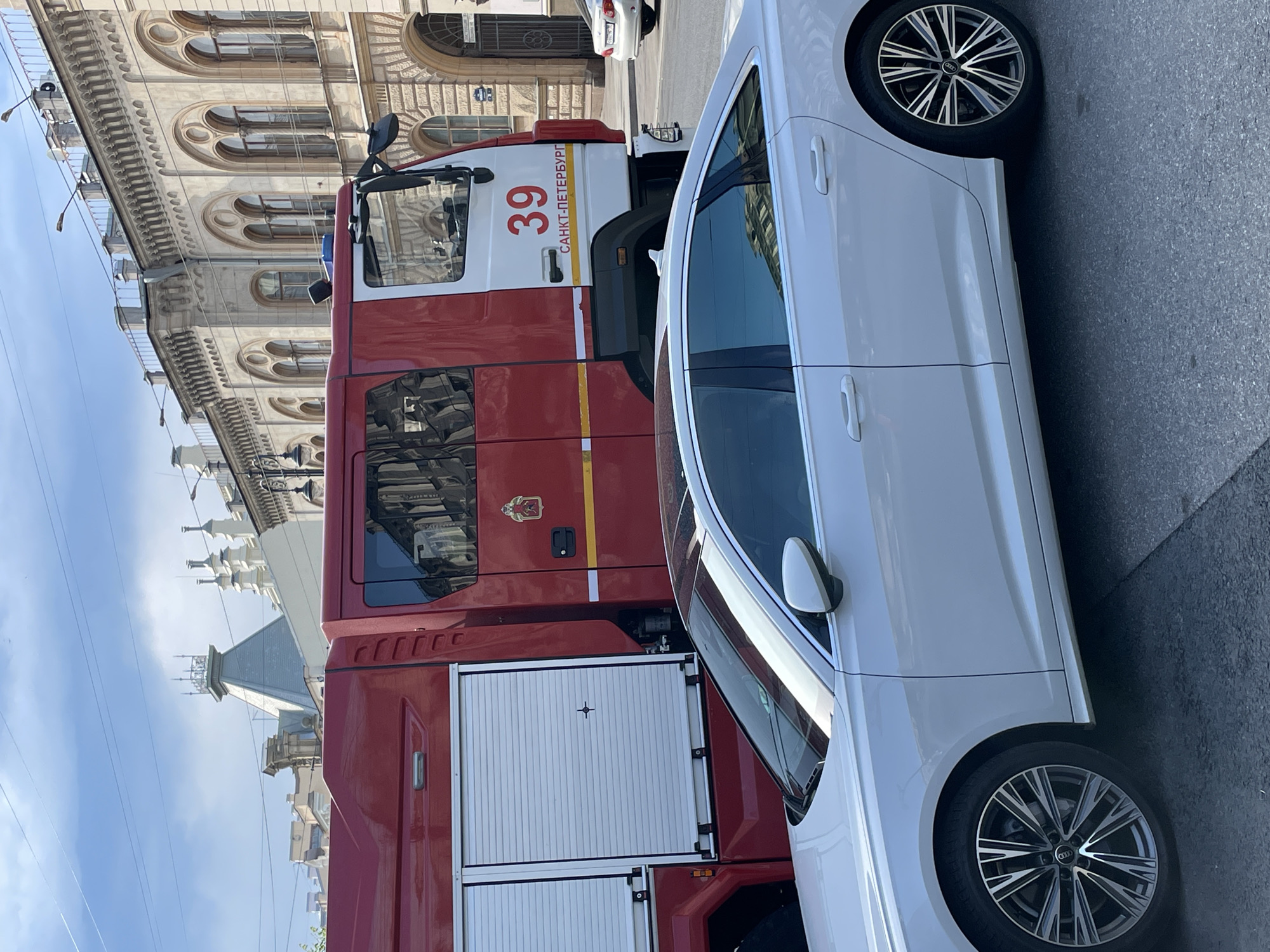 The image captures a scene on a street in Saint Petersburg, Russia. A red and white fire truck, marked with the number "39" and the inscription "САНКТ-ПЕТЕРБУРГ" (Saint Petersburg), is positioned adjacent to a white sedan. The architecture in the background features ornate buildings with arched windows, typical of the city's historical style. The environment appears to be a bright, sunny day. The vehicles are on the street.