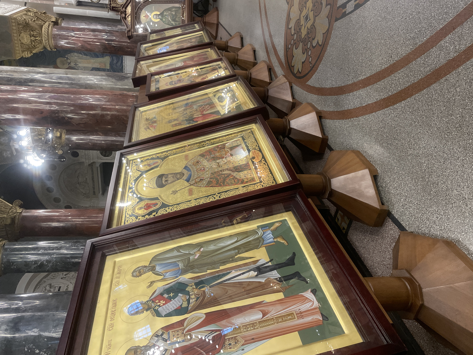 The image shows a row of framed icons displayed on wooden stands inside a church. The icons are gilded and depict religious figures in traditional Byzantine style. The church interior features large marble columns and ornate architectural details. The floor is a mosaic of patterned tiles. This scene is located in the Saint Nicholas Naval Cathedral in Kronstadt, Russia, a prominent landmark known for its religious significance and grandeur.