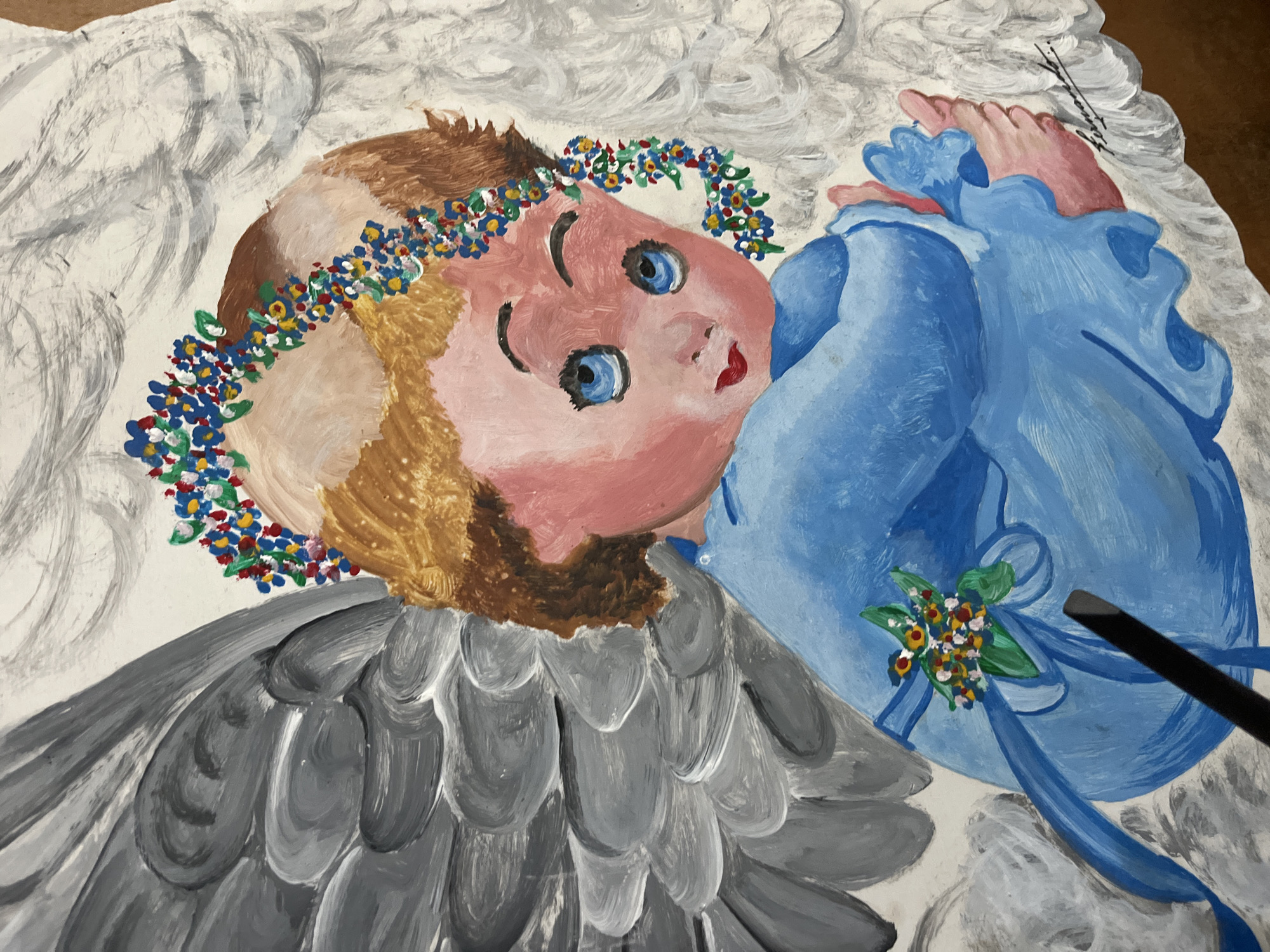 An eye-level, medium shot of an amateur painting of a blonde, fair-skinned baby or cherub with blue eyes wearing a flower crown. The baby is dressed in a blue gown with matching ribbon. The baby is lying on its back with its arms crossed across its chest, with its feet near its head. The angel has white wings and gray shoulder feathers, but the feathers do not appear to go all the way down the body. The painting is presumably in Juan Griego, Venezuela.