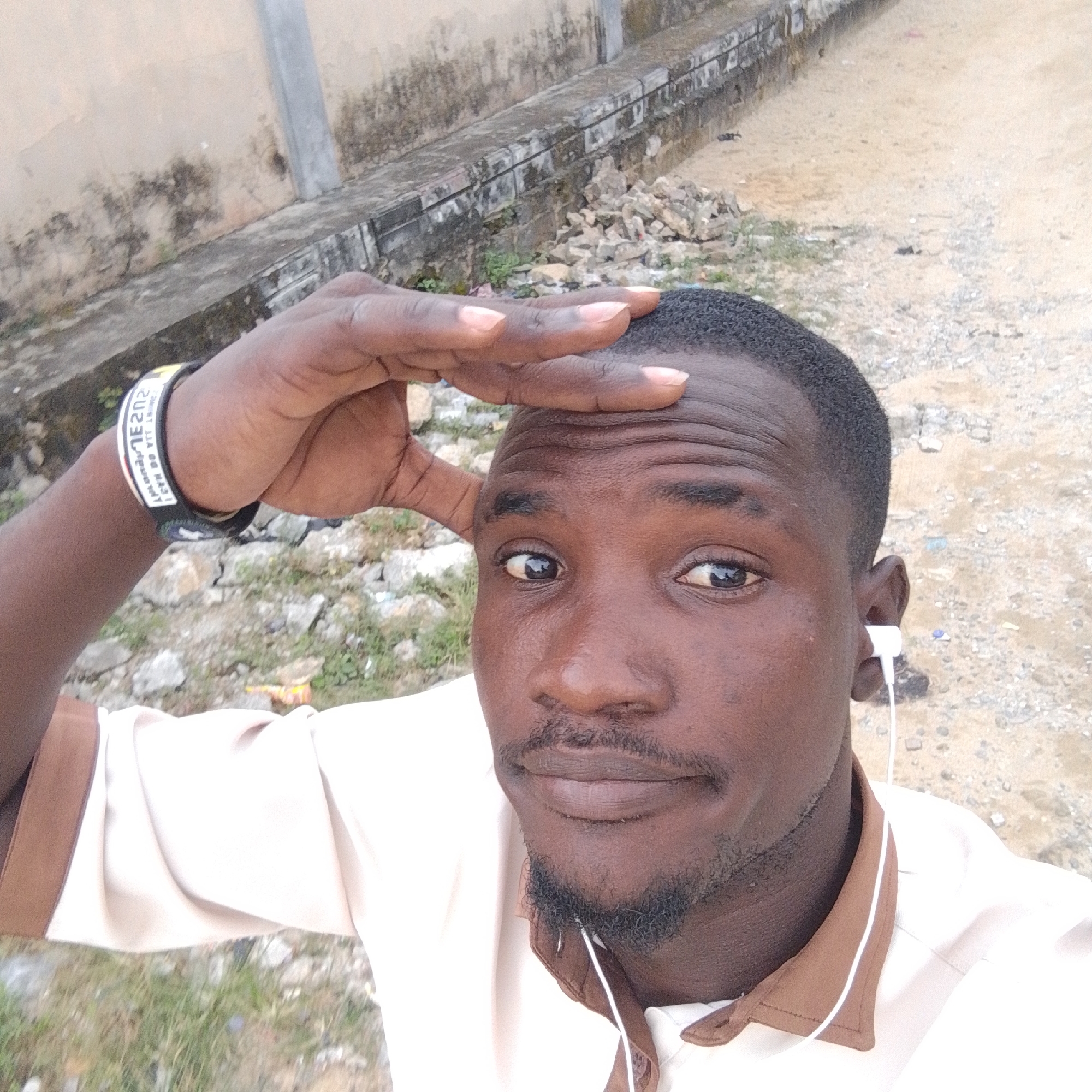 A close-up, selfie-style portrait of a Black man is captured outdoors in Ado, Nigeria. The man, appearing to be in his late twenties to early thirties, is looking directly at the camera. He has short, dark, curly hair, dark brown eyes, and a light mustache and goatee. A white earbud is visible in his right ear. He is wearing a light-colored shirt with a brown collar, and a black and yellow wristband is on his left arm. His right hand is raised, with his fingers touching his forehead as if shielding his eyes from the sun or in a gesture of contemplation.
The background suggests a somewhat unkempt outdoor environment. To the left, a weathered, light-colored concrete wall with dark stains runs horizontally, bordered by a raised concrete ledge. Beyond the ledge, the ground is a mix of dirt, gravel, and sparse green vegetation. In the distance, the ground appears to be a dirt path or road. The lighting indicates it is daytime, with harsh light casting some shadows. The overall impression is a candid moment captured in an everyday setting within Ado, Nigeria.