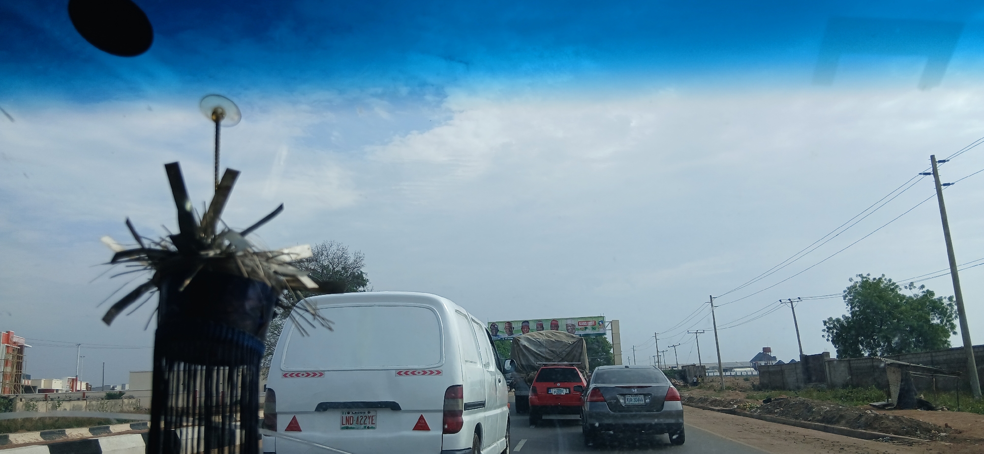 The image is taken from inside a vehicle looking out onto a road with several other vehicles in Tudun Wada, Nigeria. A white van is in front with the license plate LND-422YE. There is a red vehicle and a gray vehicle further ahead. A billboard advertising political candidates is in the background. Electrical poles line the side of the road. Debris and garbage are along the edge of the road. A decorative object hangs from the rearview mirror of the vehicle the image was taken from.