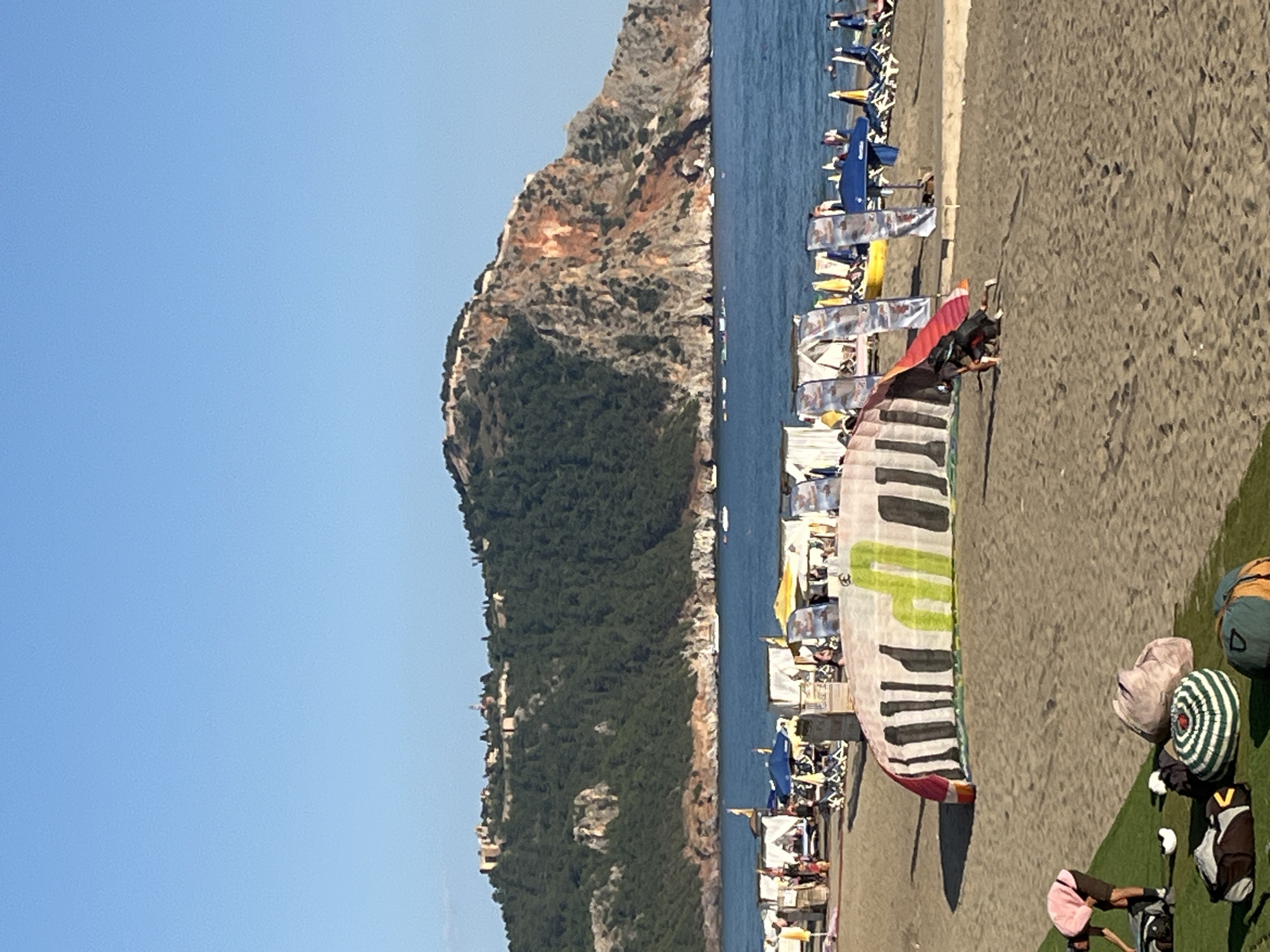 The image shows a scene on a beach in Alanya, Turkey, under a clear blue sky. A rocky mountain covered in vegetation is visible along the coast. The beach is lined with beach chairs and umbrellas. In the foreground, a person is setting up a paragliding kite with a design resembling a piano keyboard. Golf clubs and bags rest on the green strip to the right side of the image. People are present on the beach, but their activities are not clear due to the distance. The sea appears calm.